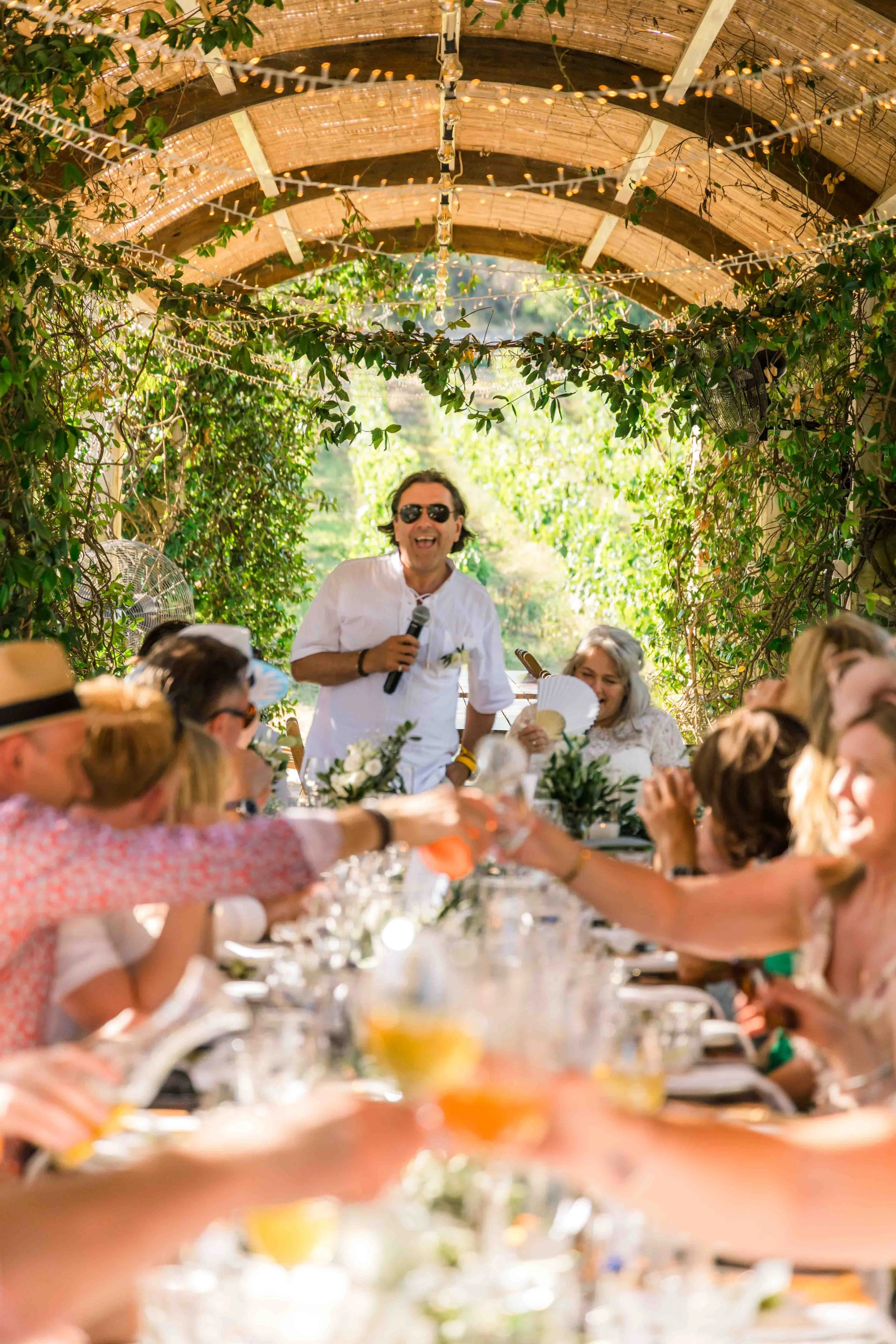 A man in sunglasses standing and holding a microphone at a long outdoor dining event with string lights and lush greenery.