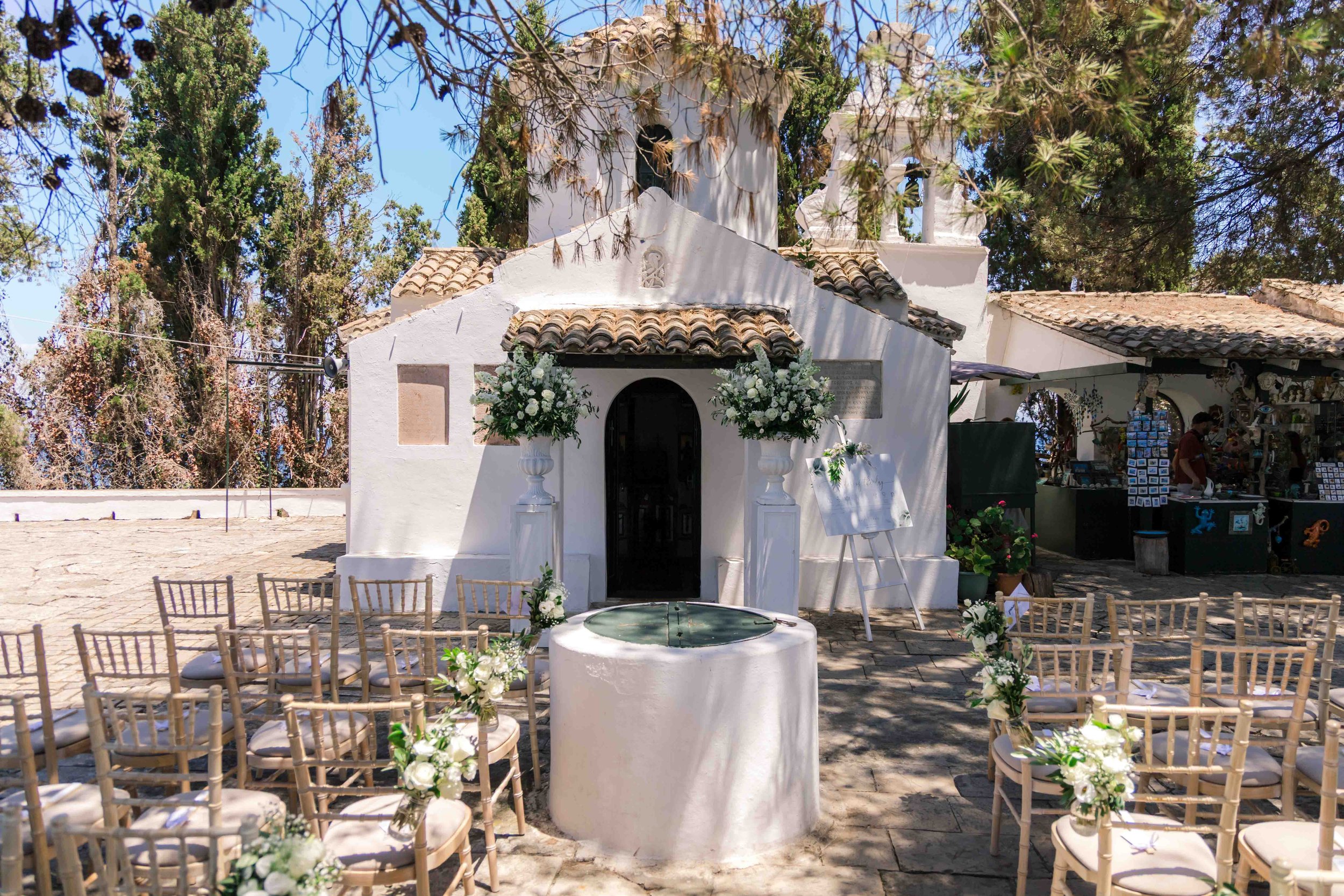 Small white chapel decorated with white flowers and greenery, set up for an outdoor wedding ceremony with chairs arranged on either side of the aisle, and a fountain in front of the entrance under a tree canopy.