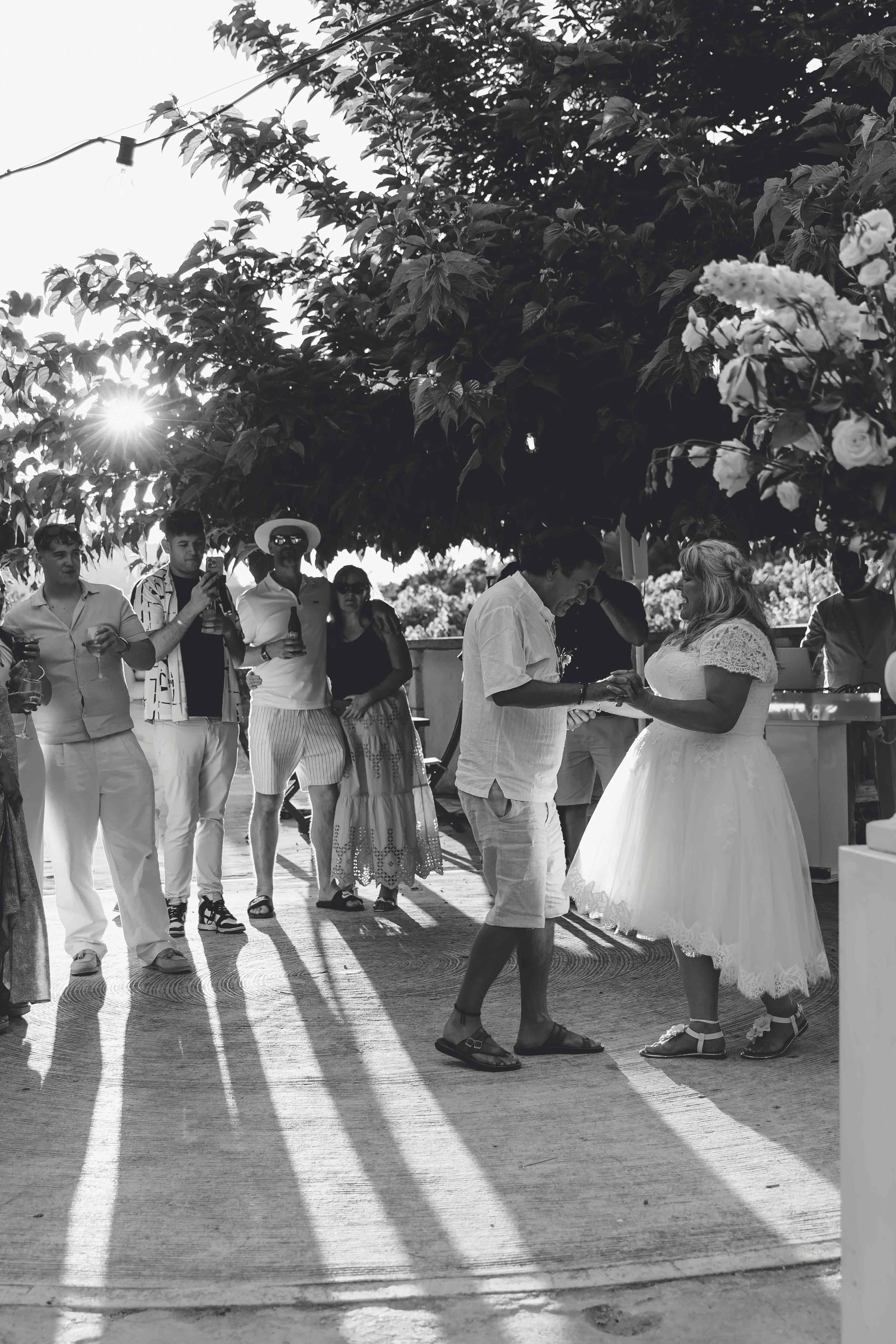 A group of people at a wedding, with some taking photos, standing underneath a tree with sunlight shining through the leaves. The bride and groom are in the foreground, holding hands, with the bride in a white wedding dress and the groom in casual at