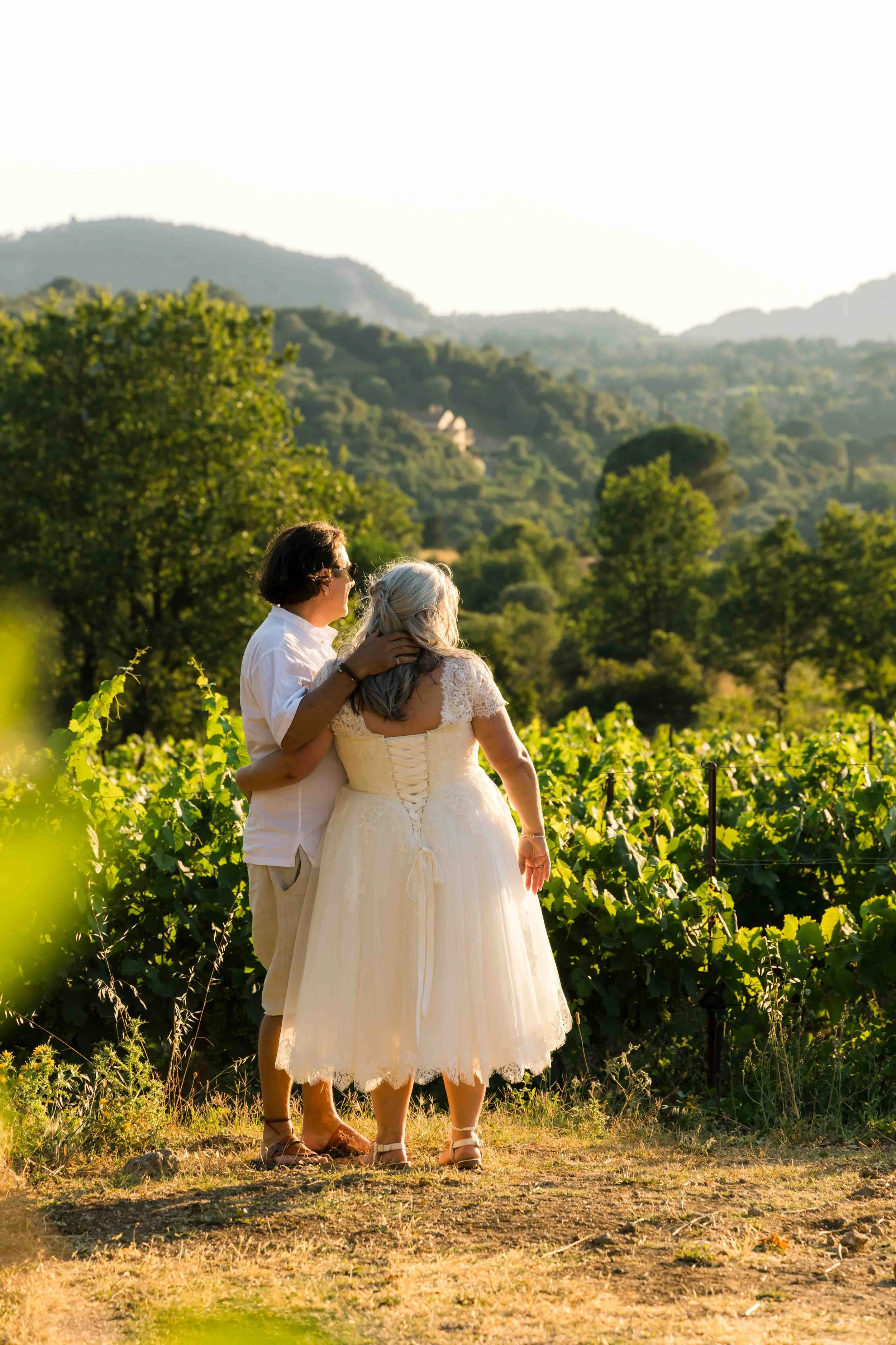 A couple dressed in light clothing, with the woman in a wedding dress, standing in a vineyard during sunset, with green hills and mountains in the background