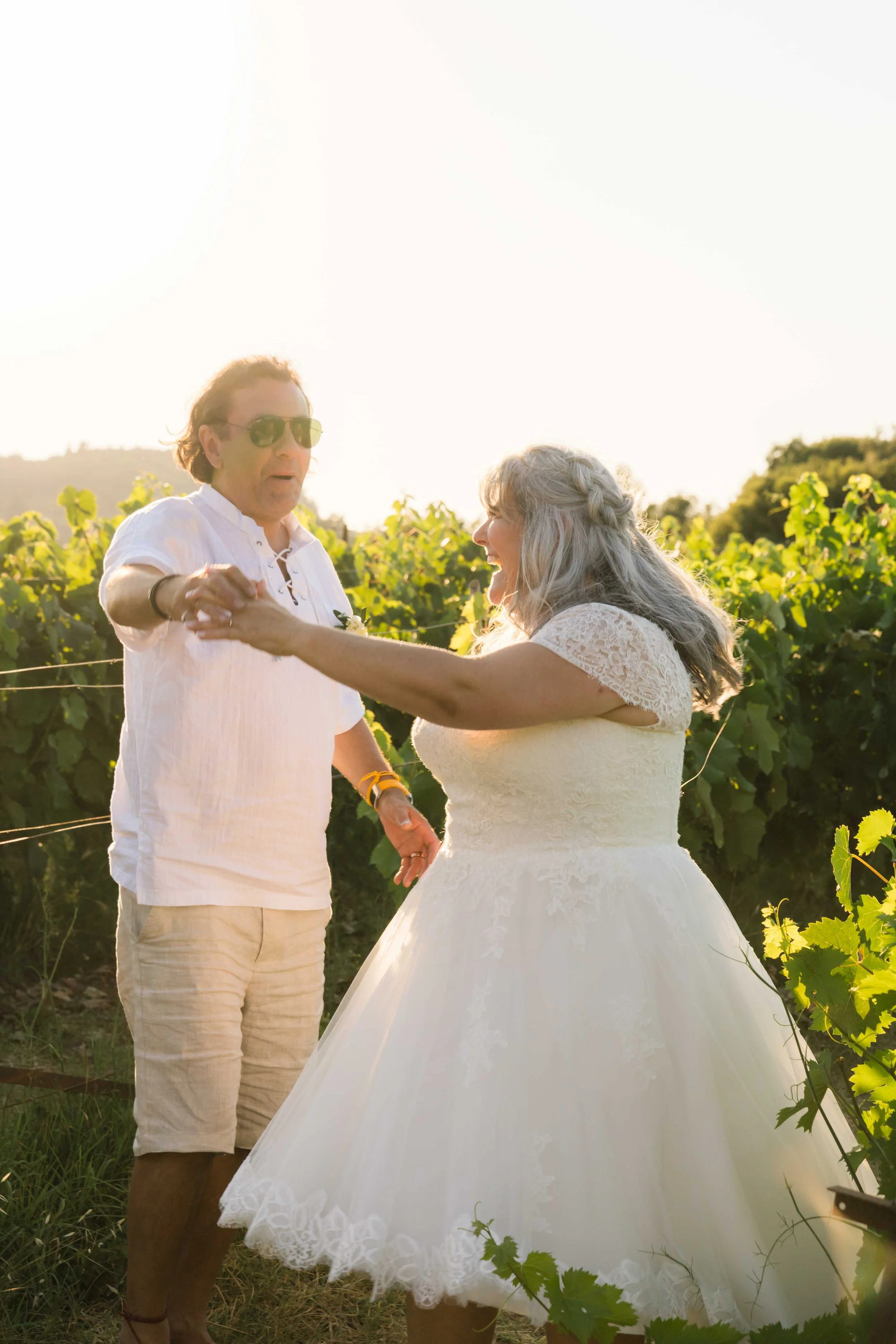 A man and woman are dancing together in a vineyard during sunset. The woman is wearing a white wedding dress, and the man is in casual summer clothes with sunglasses. They are holding hands and smiling.