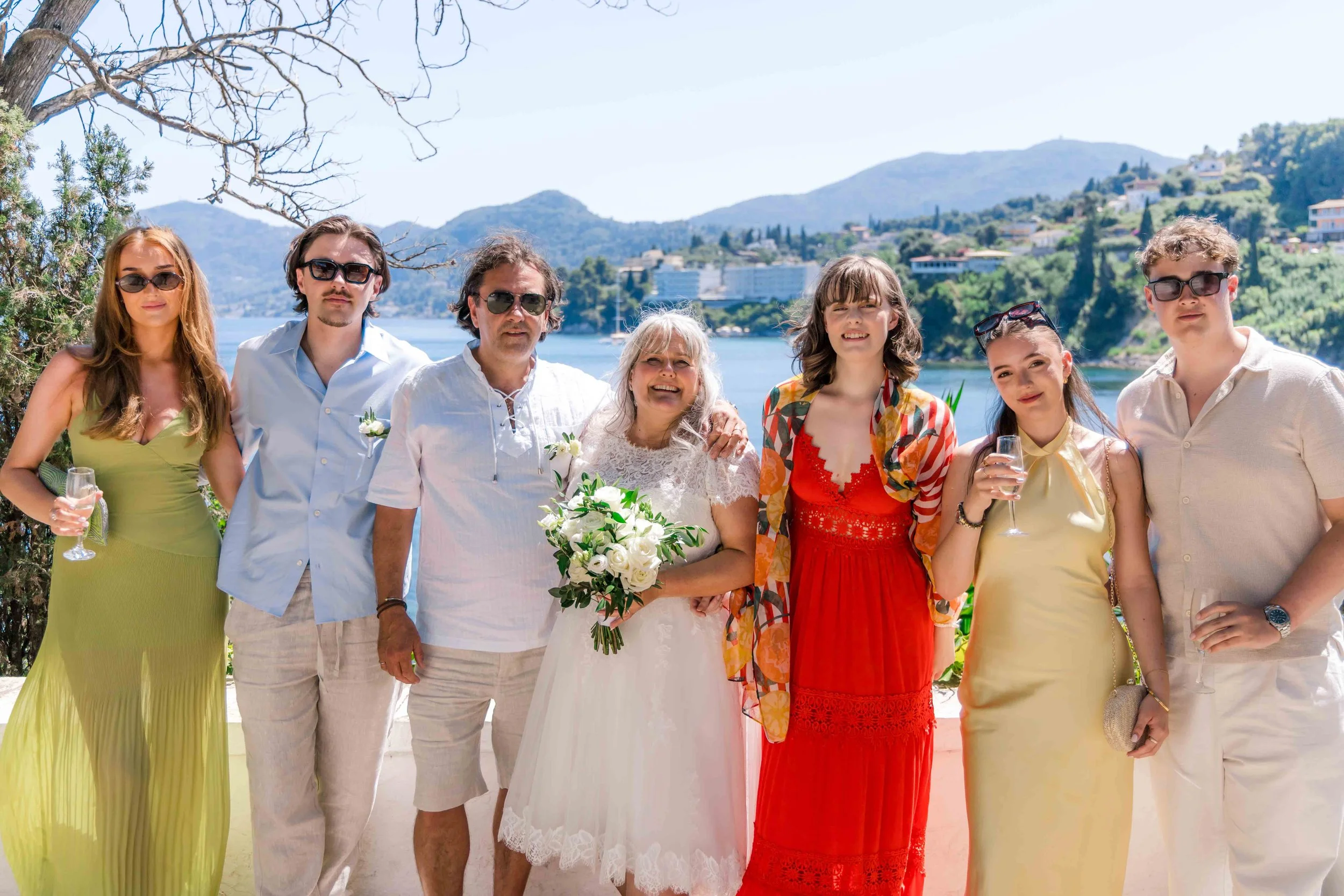Group of eight people dressed in summer clothes standing outdoors near a lake with mountains in the background, celebrating a wedding.