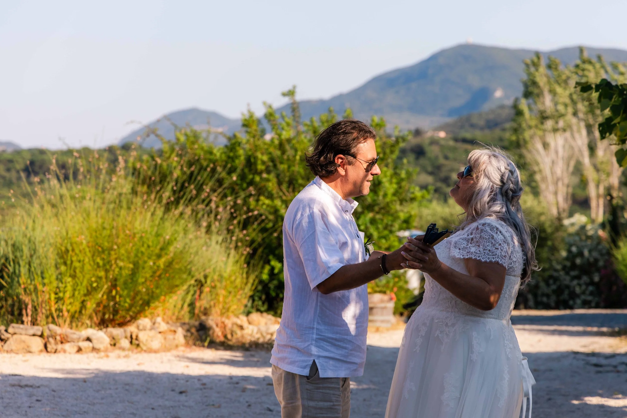 A man and woman exchanging wedding vows outdoors, holding hands, with a mountainous landscape and clear sky in the background.
