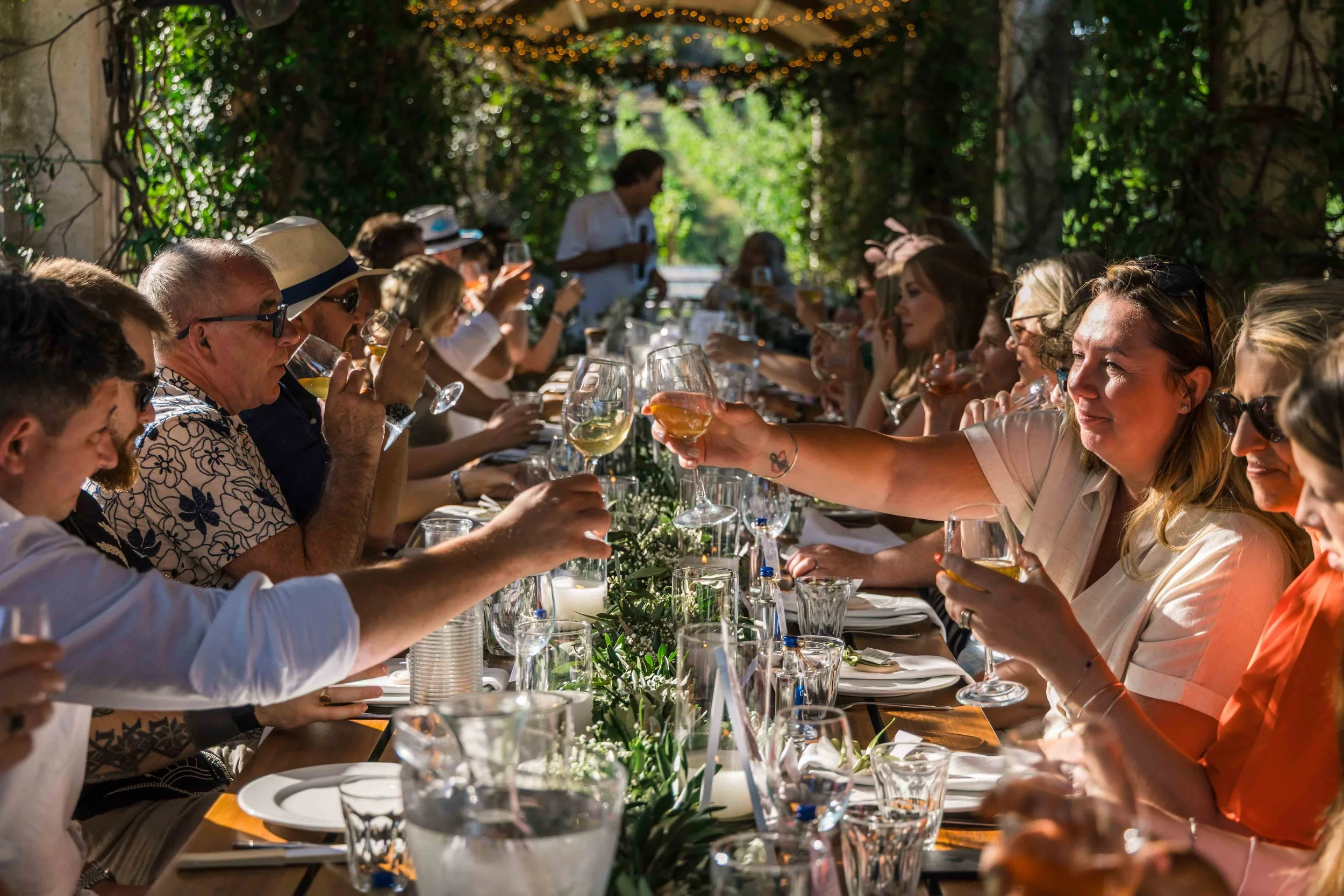 People sitting at a long outdoor dining table with glasses of wine and food, surrounded by greenery and string lights, enjoying a social gathering.