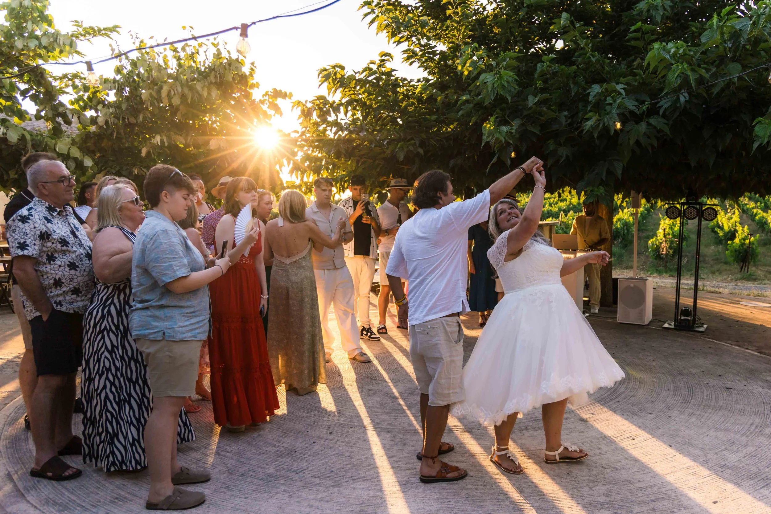 A bride in a white wedding gown is dancing with a man at an outdoor wedding reception during sunset. A group of guests, some taking photos, watch and enjoy the moment, surrounded by lush greenery and warm sunlight.