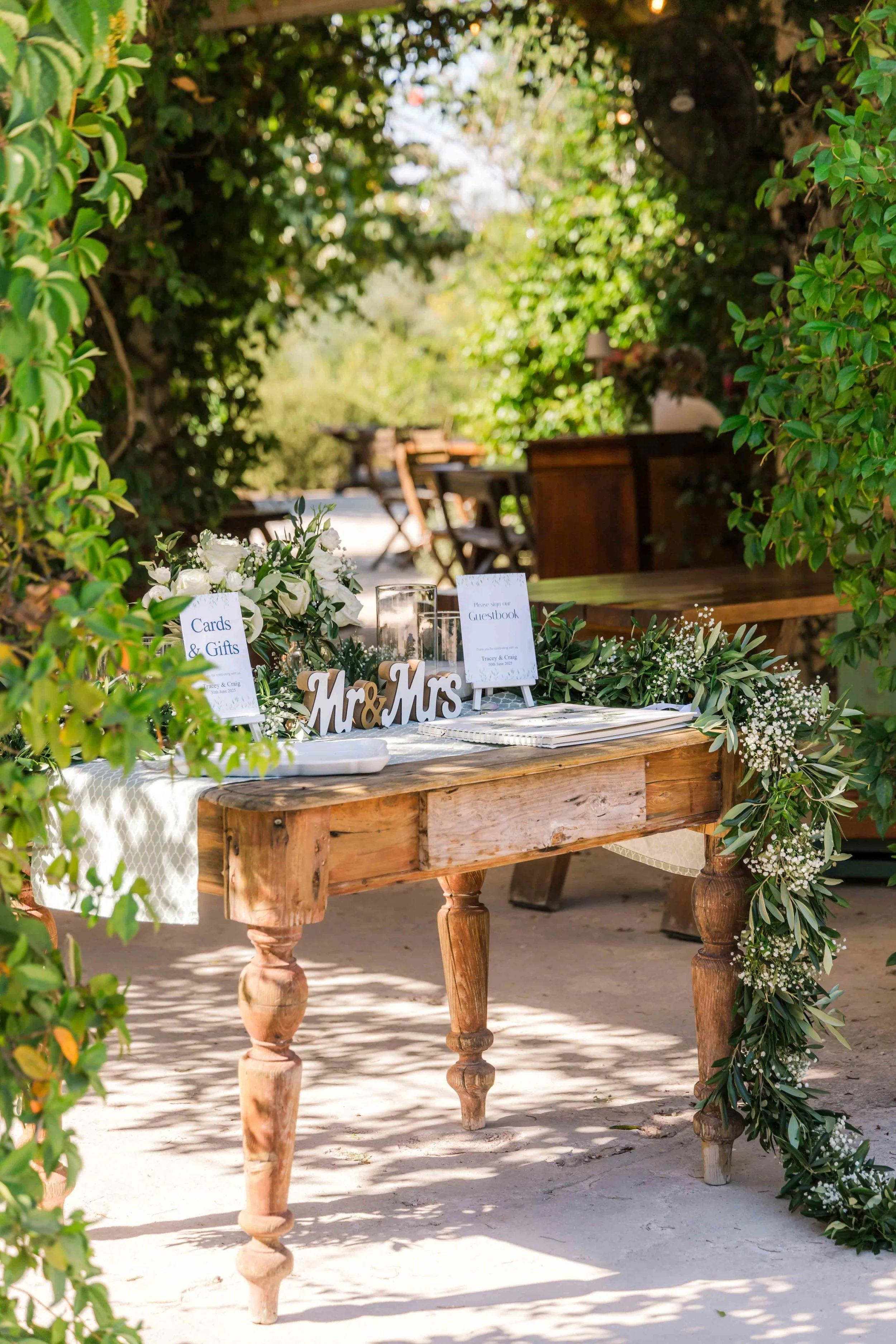 Outdoor wedding reception area with a rustic wooden table decorated with fresh white flowers, greenery, and 