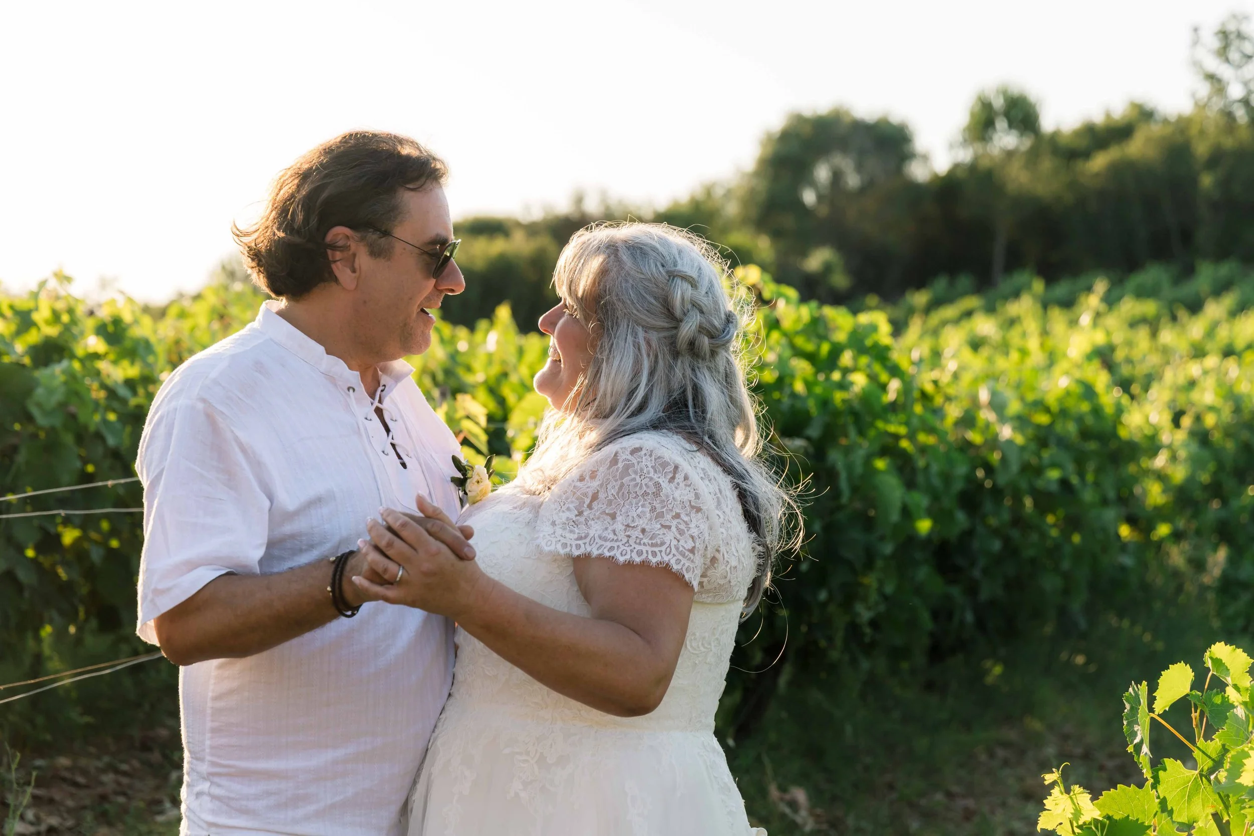 A couple dancing outdoors in a vineyard during sunset, holding hands and looking at each other, with green grapevines in the background.