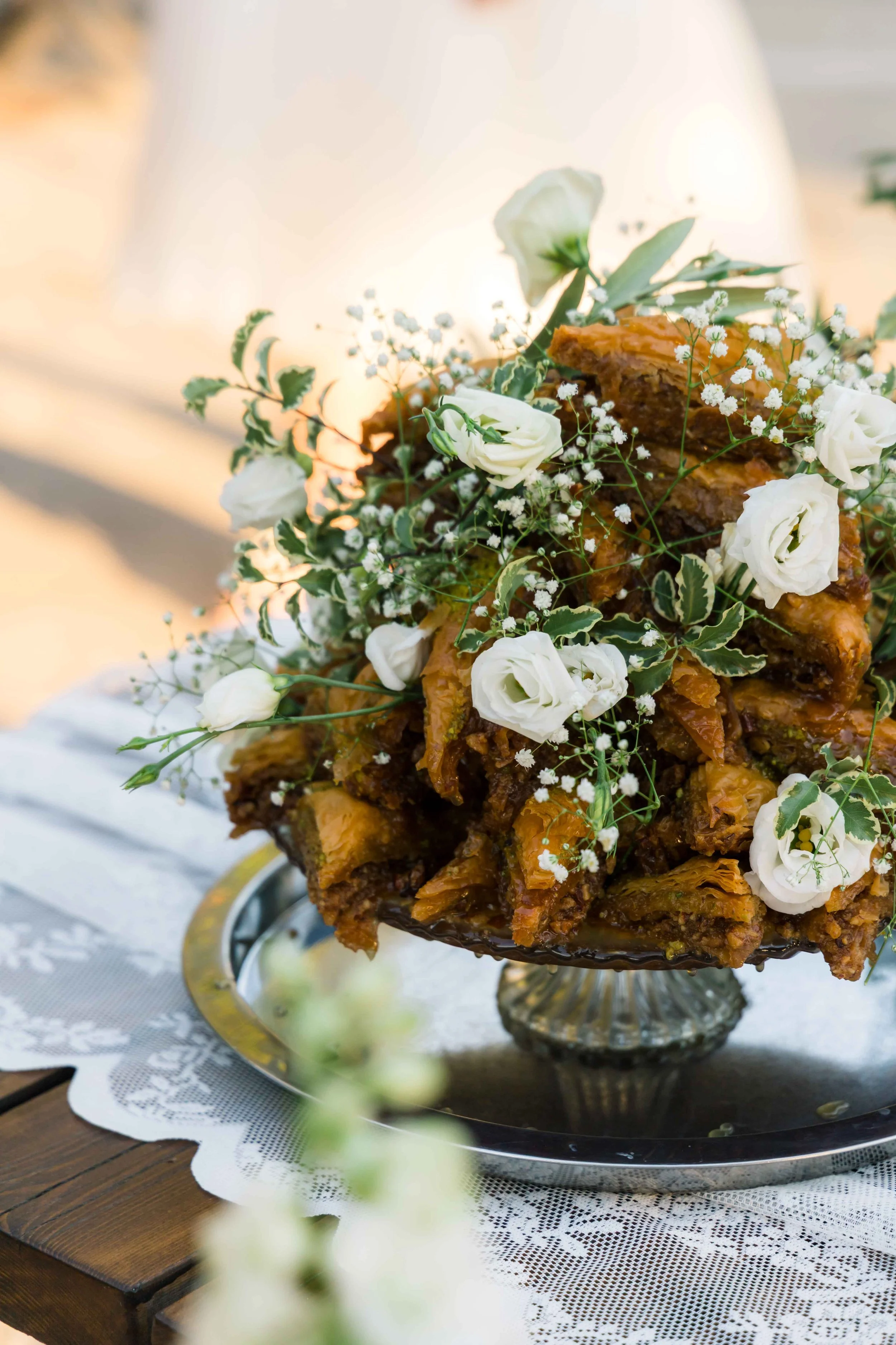 A pie topped with white roses and baby's breath flowers, displayed on a silver pedestal on a lace tablecloth.
