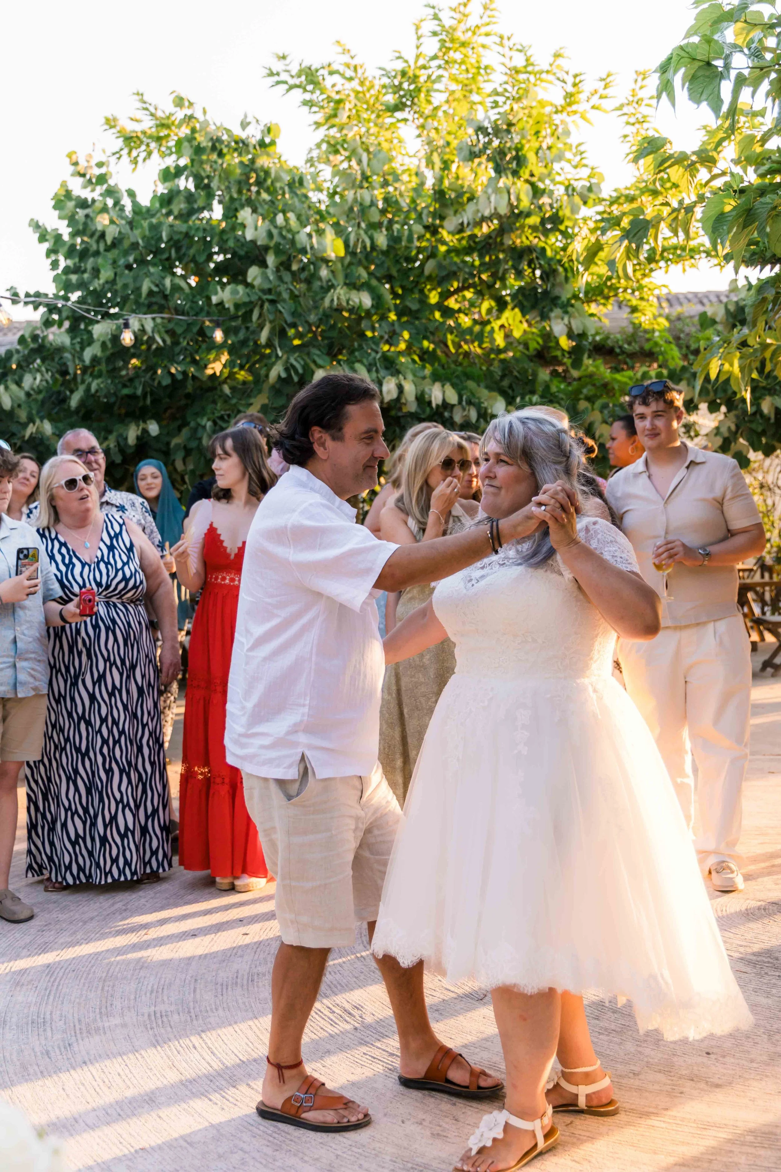 A couple is dancing at an outdoor wedding reception surrounded by friends and family on a sunny day. The man is wearing a white shirt and beige shorts, and the woman is in a white dress with lace details. Guests are standing around, some with cameras