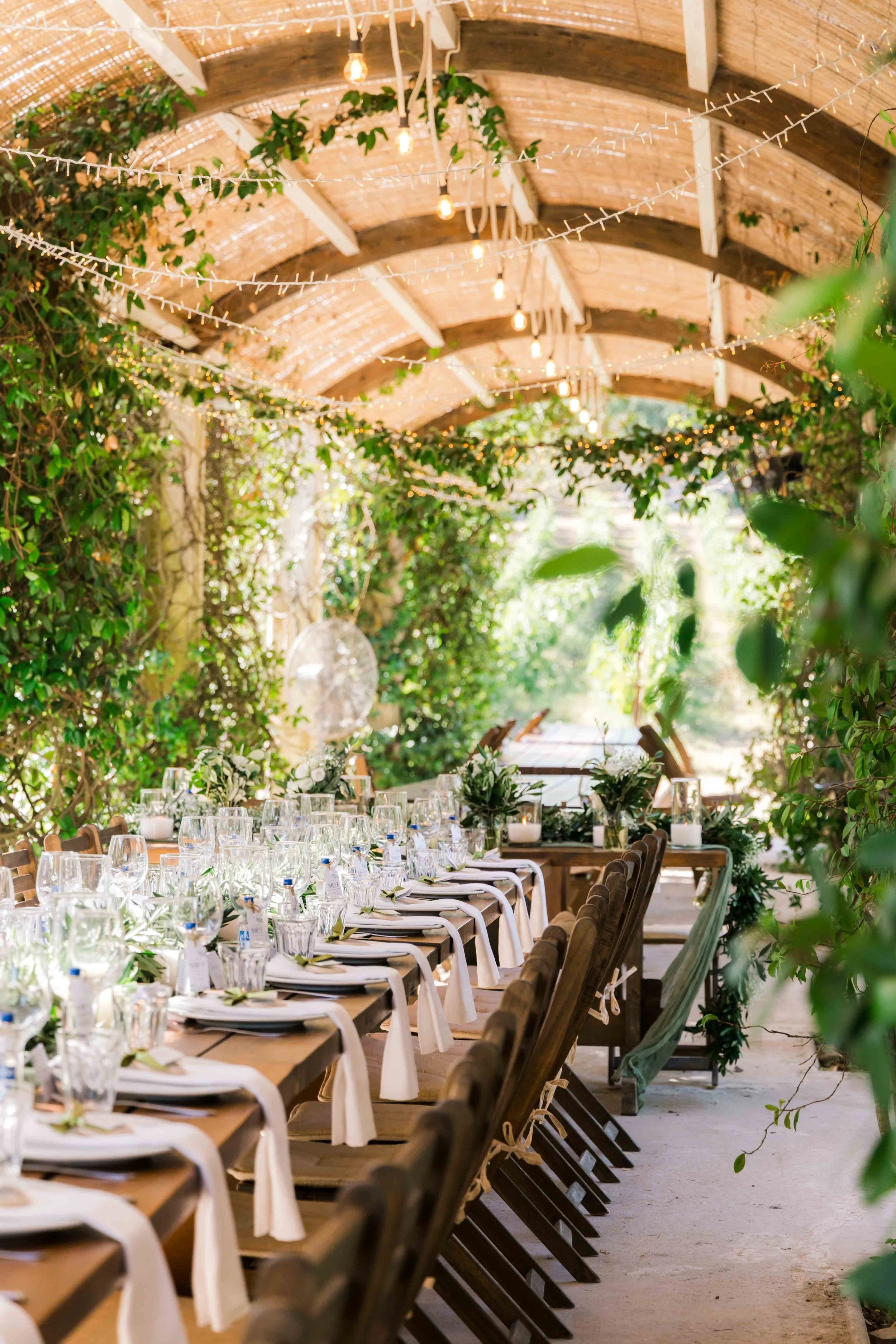 Long outdoor table set for a celebration, decorated with greenery and glassware, under a wooden pergola draped with string lights and surrounded by lush green foliage.