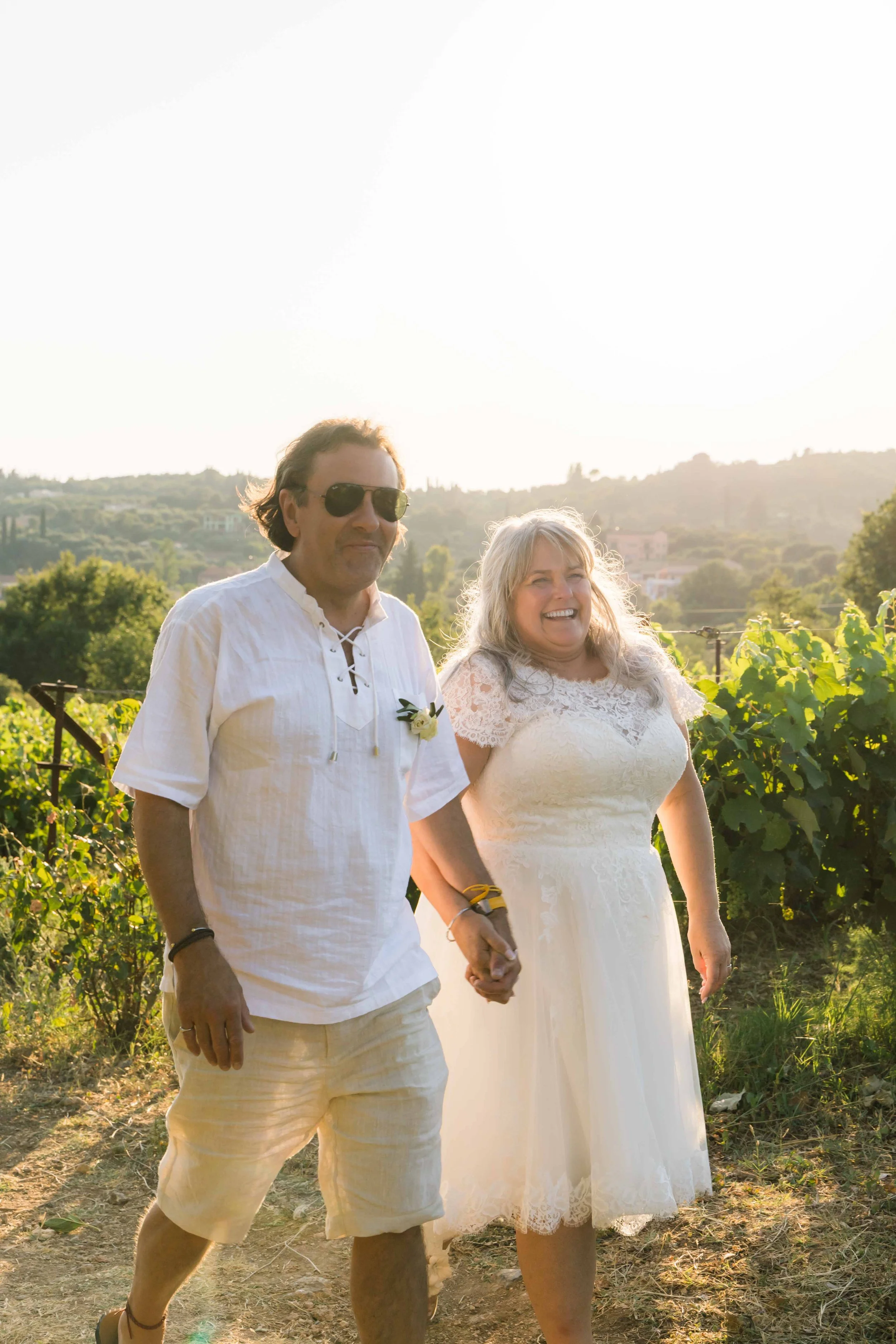 A happy couple walking hand in hand outdoors in a lush green vineyard during the day, with hills and houses in the background, and bright sunlight.