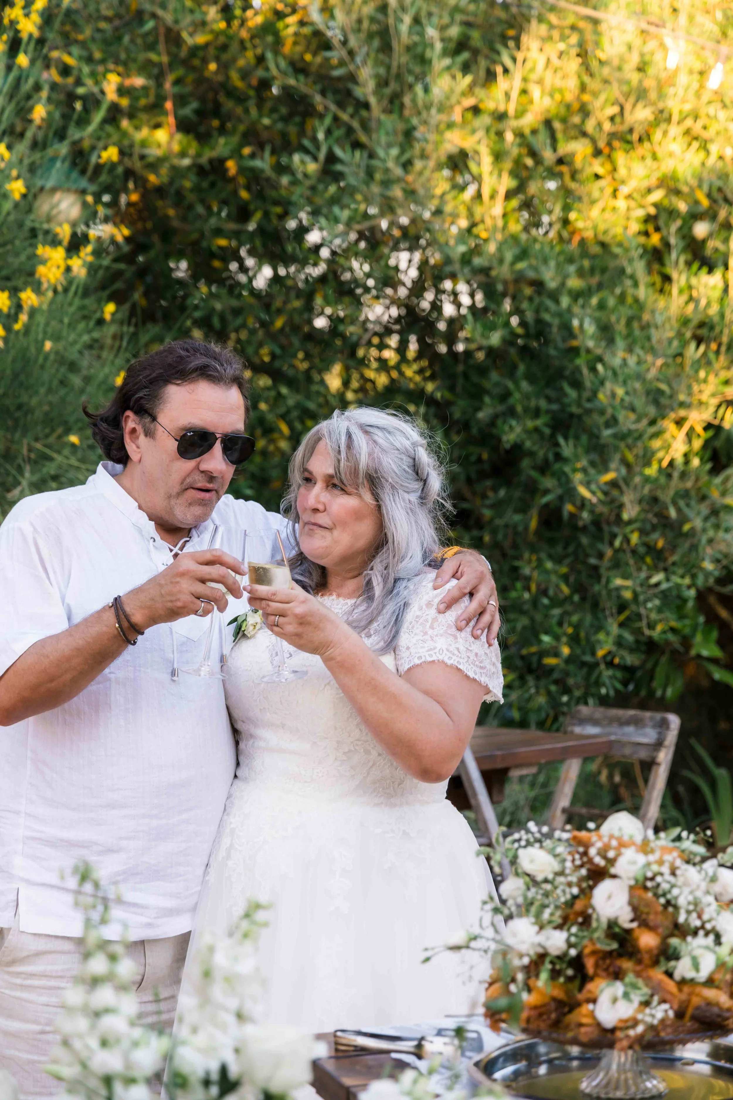 A middle-aged couple with light skin and gray hair, dressed in white, sharing a toast at a wedding reception outdoors. The man wears sunglasses and holds a champagne flute, and the woman smiles, holding a similar glass. The background features lush g