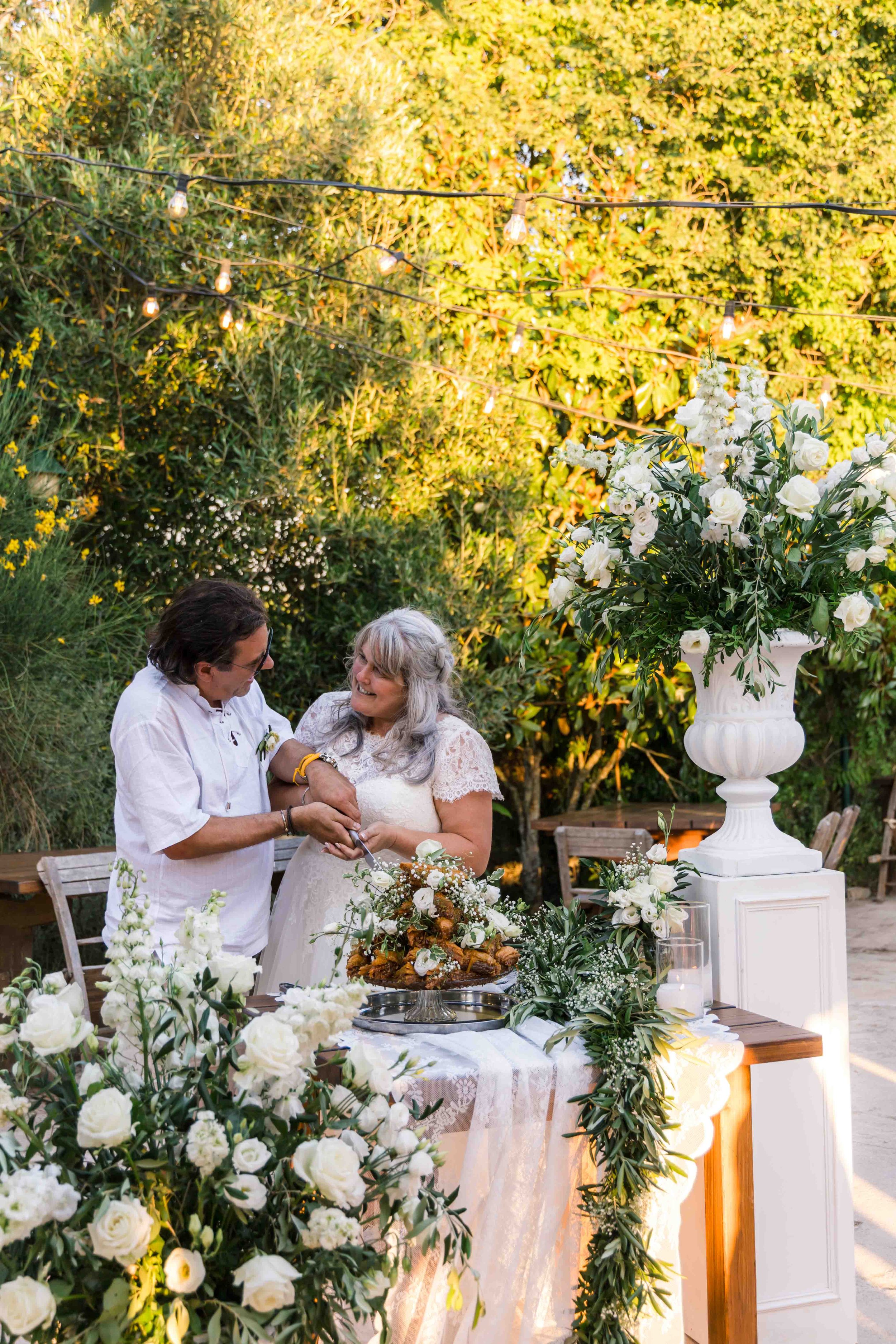 A wedding reception outdoors with a couple exchanging vows, surrounded by white flowers and greenery, with string lights overhead and a large floral arrangement on a white pedestal.