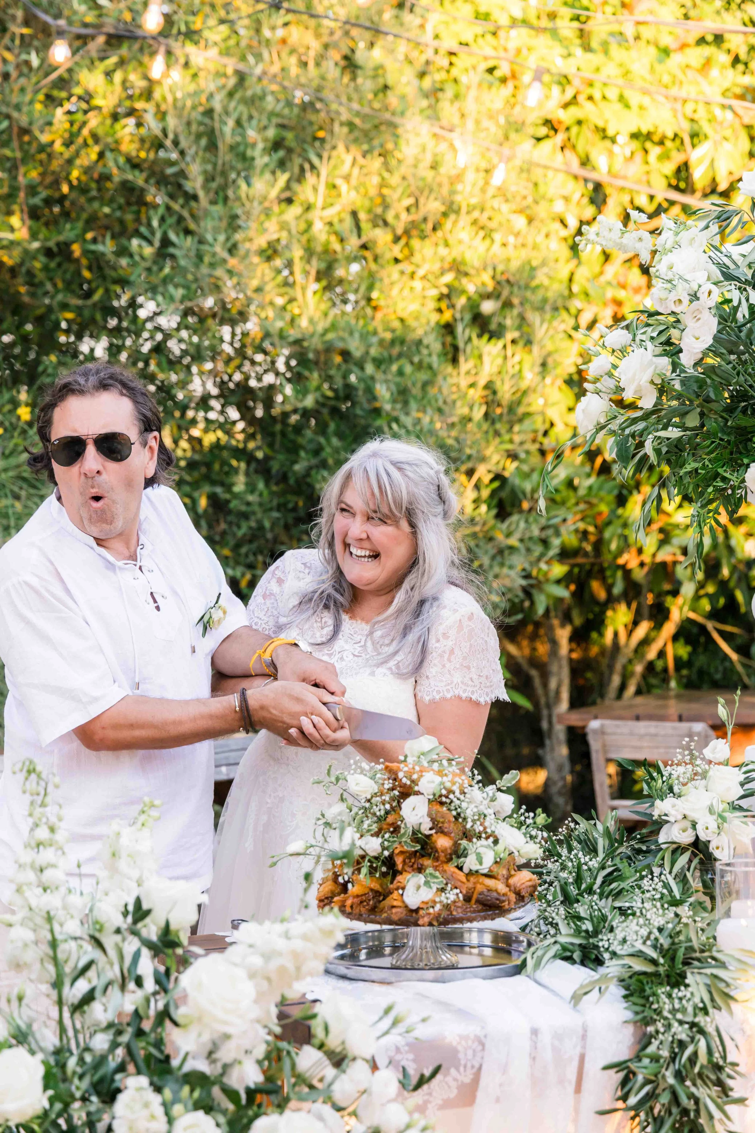 A man and a woman at a wedding reception, cutting a cake together outdoors with lush greenery and white flowers around them.