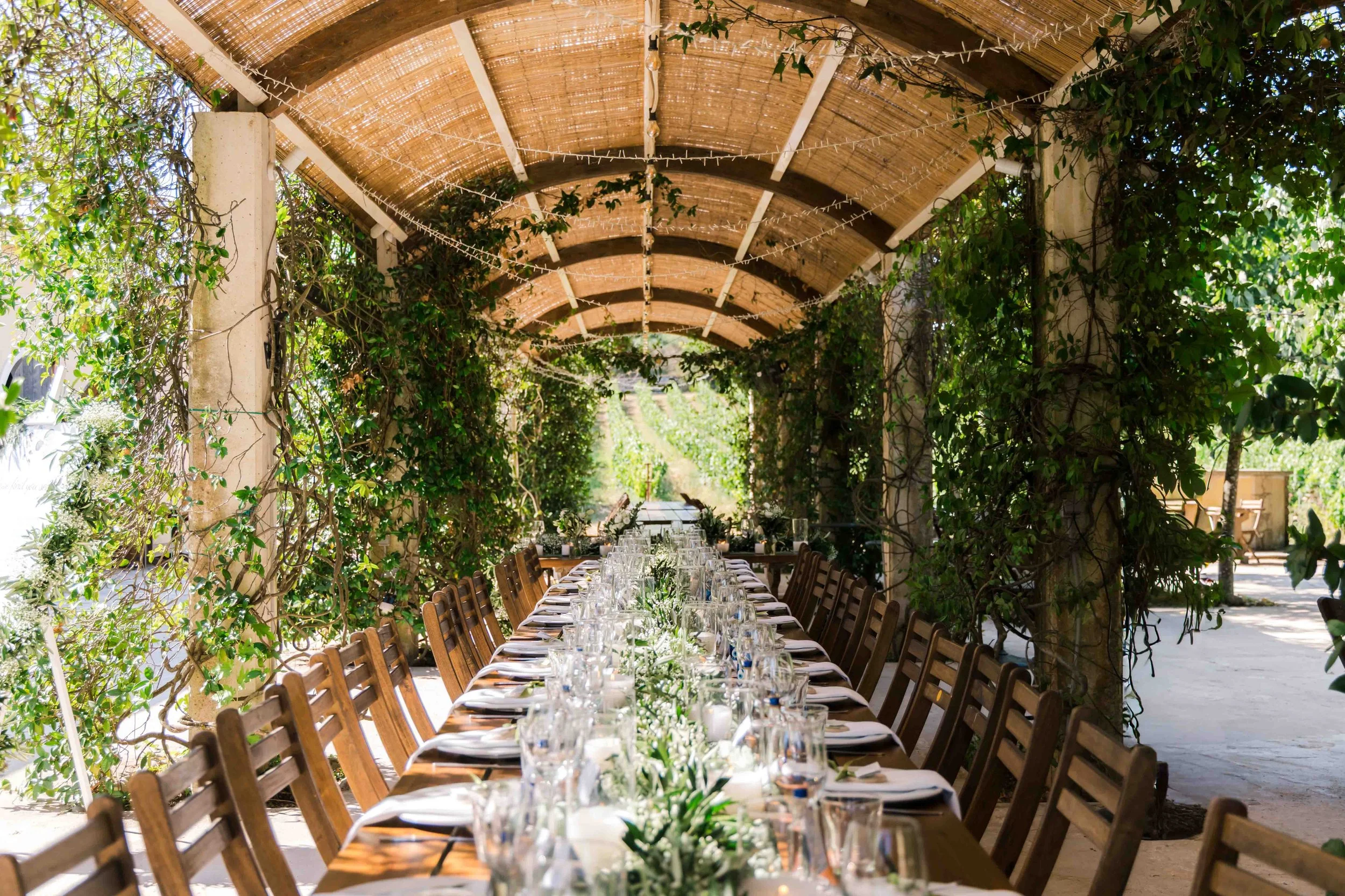 Long outdoor banquet table with white flowers, surrounded by wooden chairs, under a wooden archway with greenery and string lights in a garden setting.