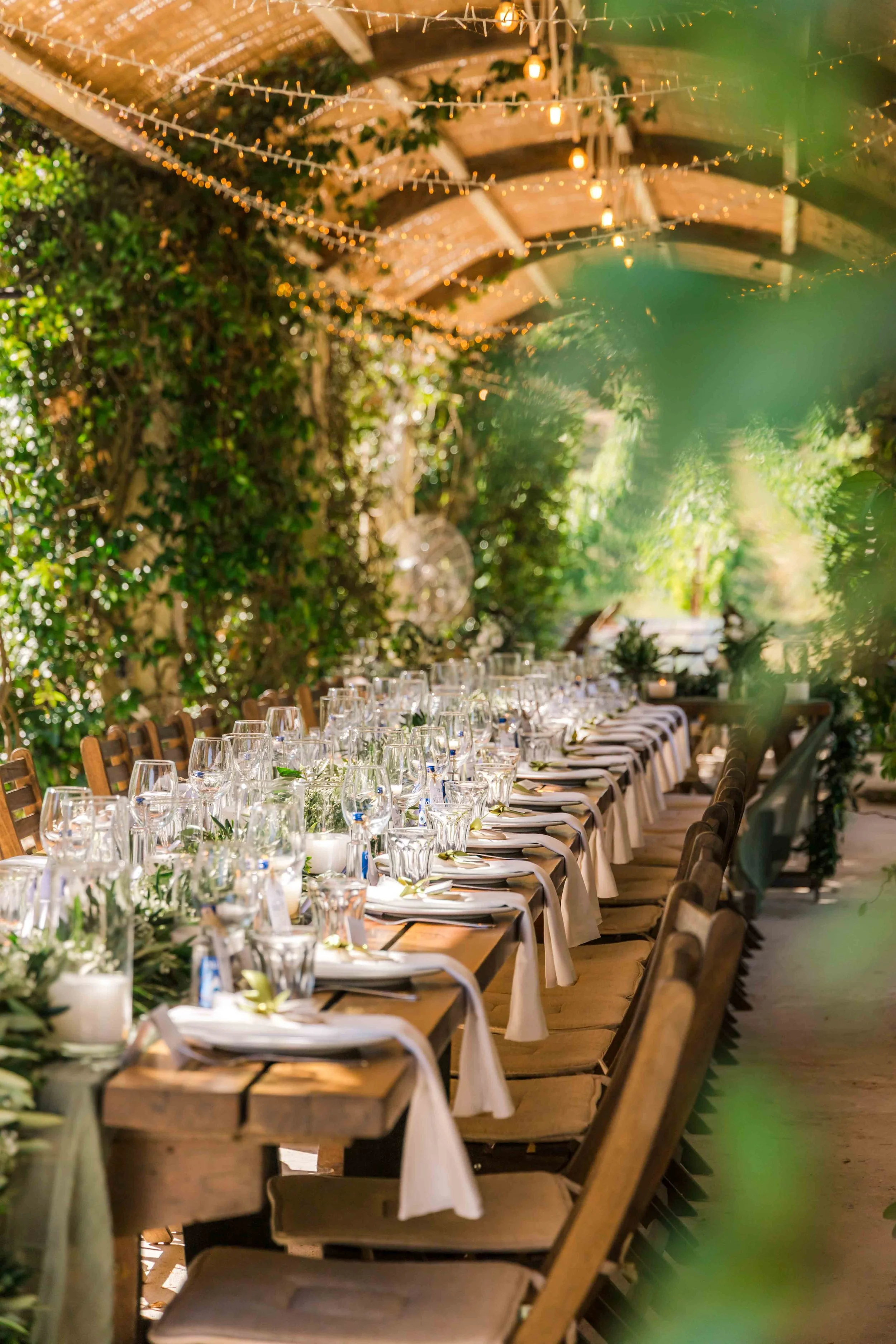 Long wooden dining table set for a formal event, decorated with white tablecloths, glassware, and greenery, under a canopy of string lights and surrounded by lush greenery.
