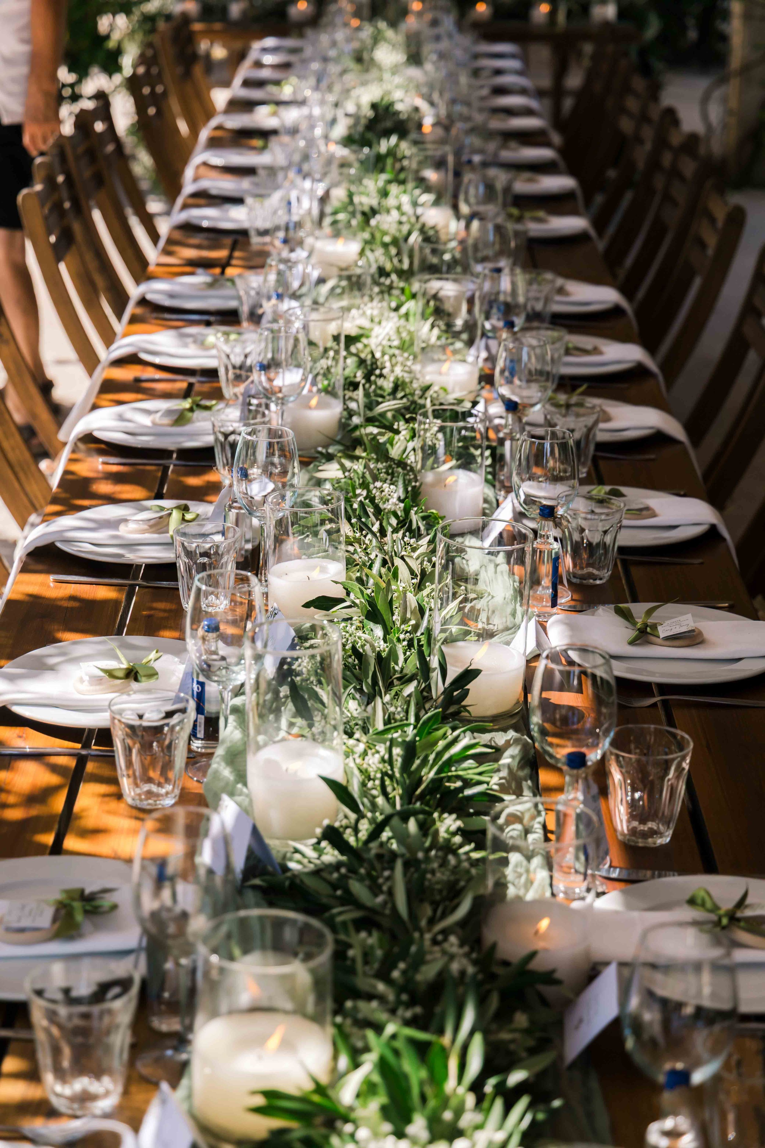 Long rectangular dining table set up for a formal event with glasses, candles, and a centerpiece of greenery and white flowers, outdoors with natural sunlight.