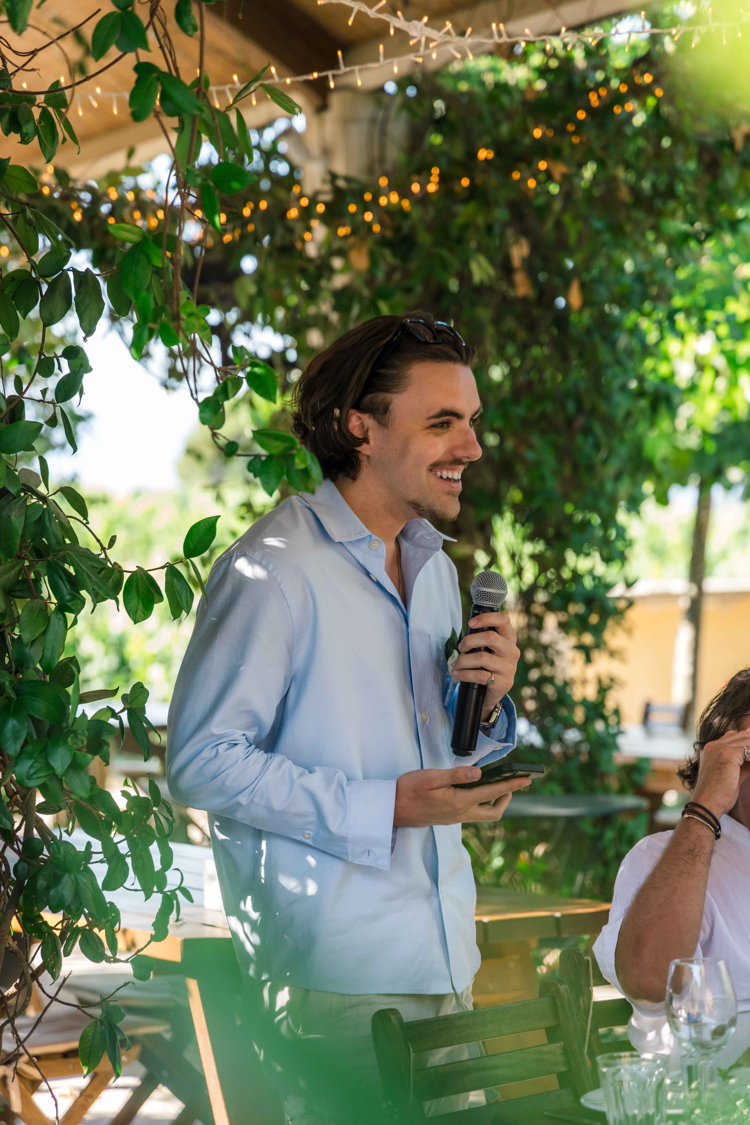 A man with dark hair wearing a light blue dress shirt smiling while holding a microphone and a phone at an outdoor event, with green foliage and string lights in the background.