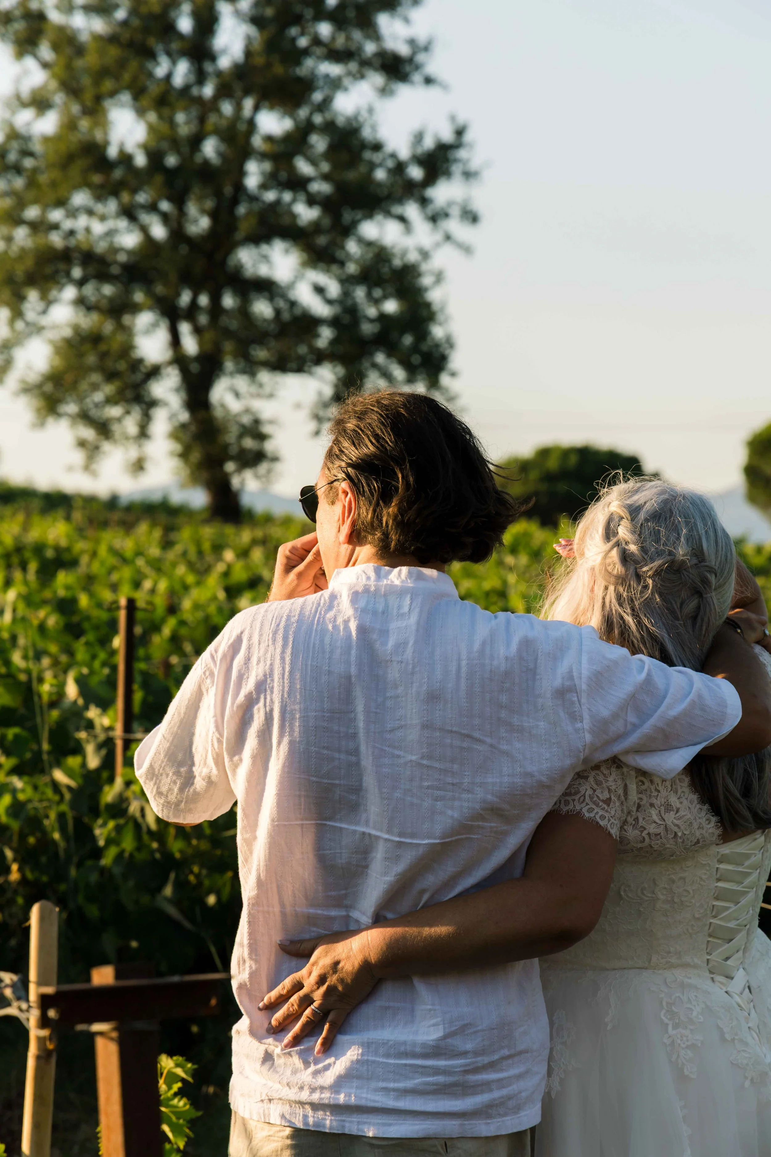 A couple hugs outdoors with the woman in a white dress with lace details and the man in a white shirt. The setting appears to be a garden during late afternoon or early evening, with greenery and a tall tree in the background.