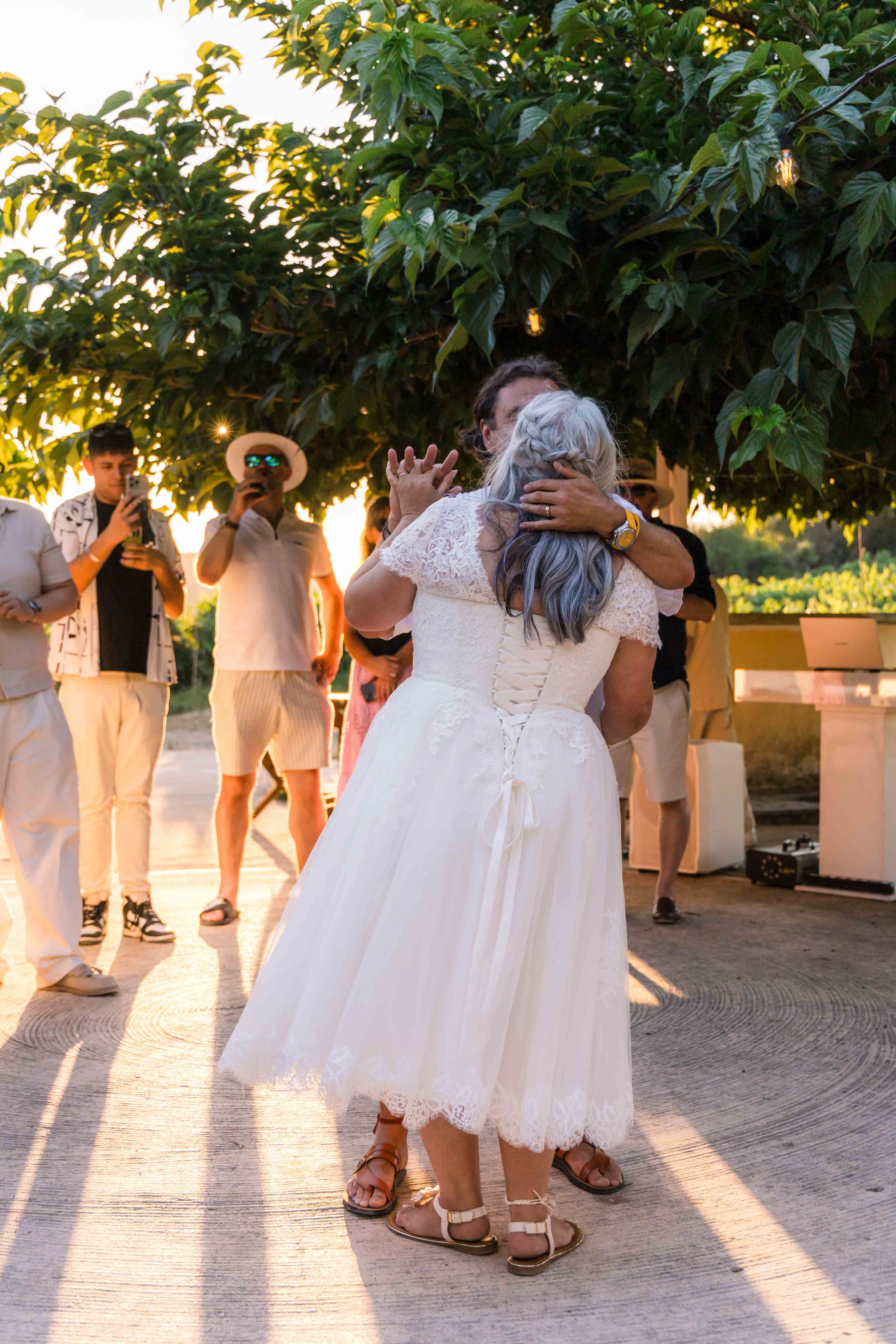 Couple dressed in white wedding attire sharing a kiss at sunset, with guests in casual summer clothing watching and taking photos in an outdoor setting with green foliage overhead.