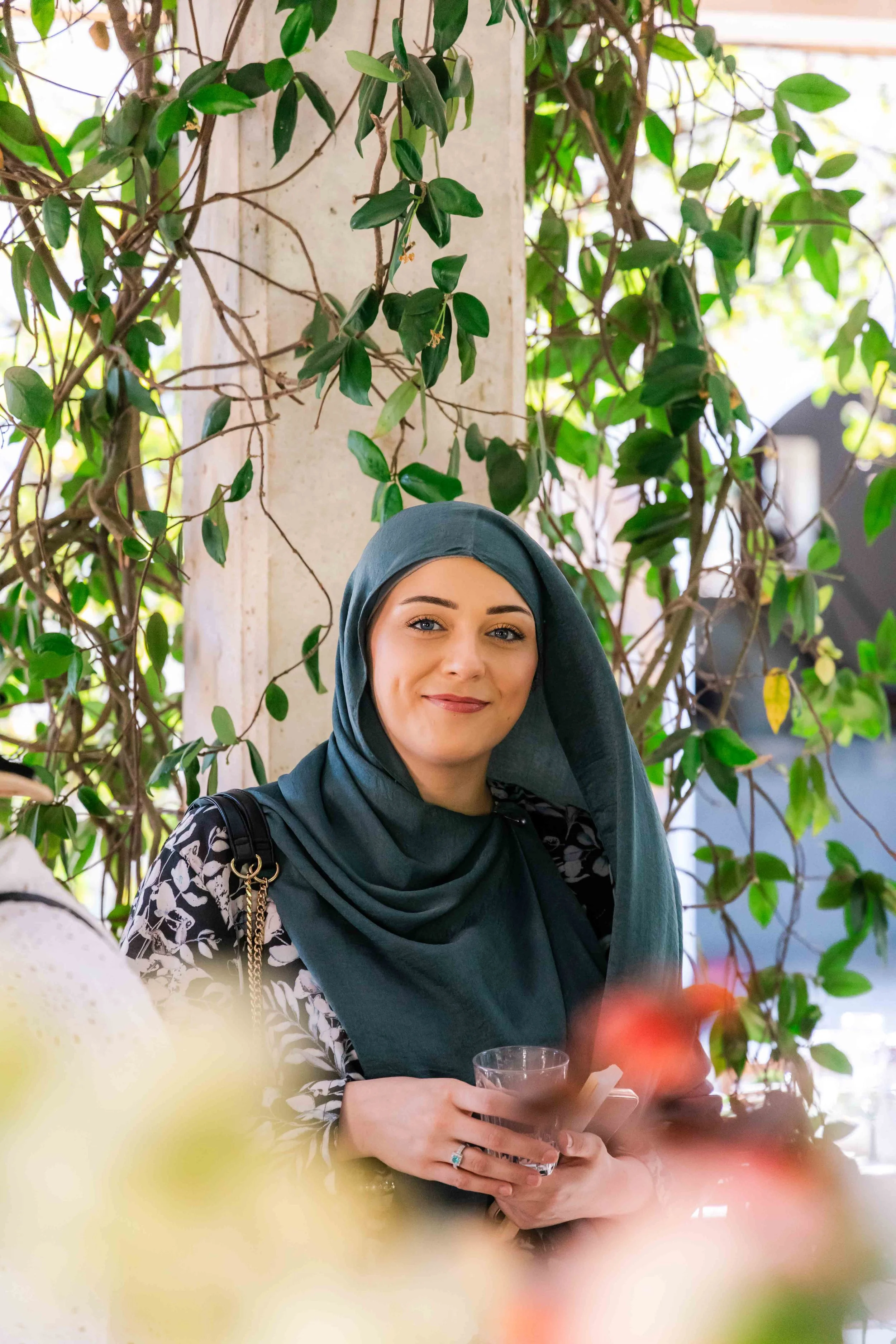 A smiling woman wearing a teal hijab, sitting at a table with a glass, surrounded by green leafy plants.