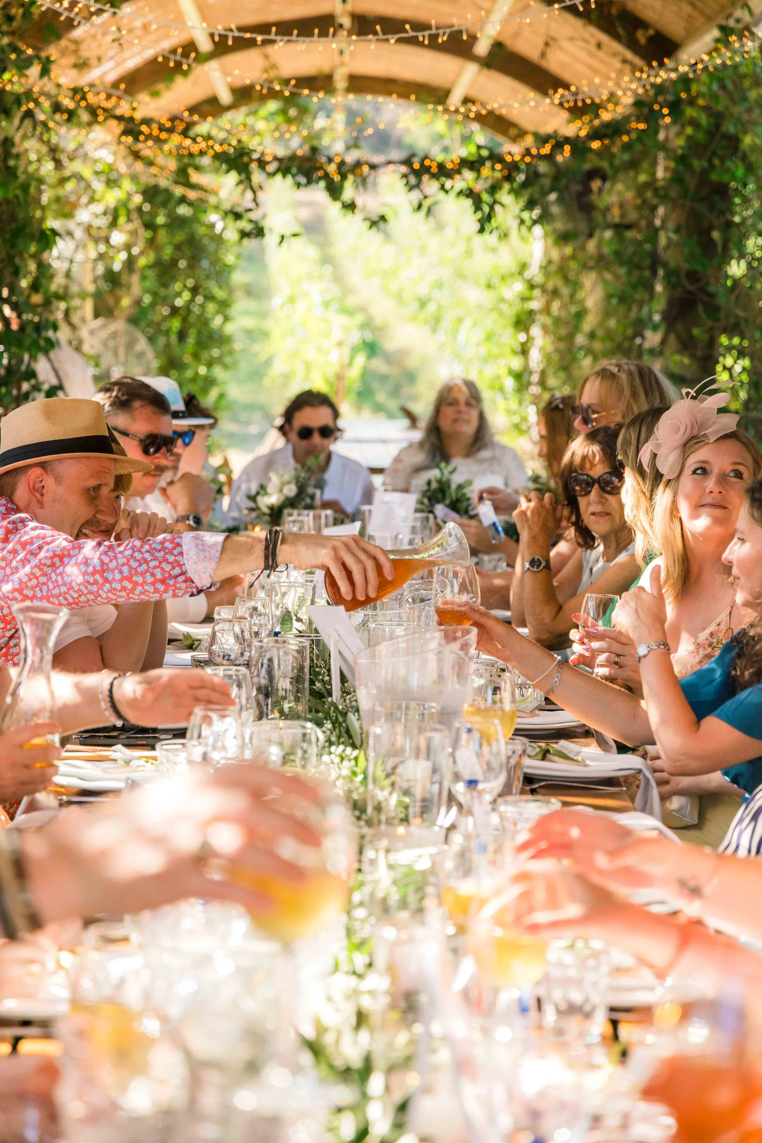 People gathered at a long outdoor table under a canopy decorated with string lights, enjoying a social meal with drinks and floral centerpieces.