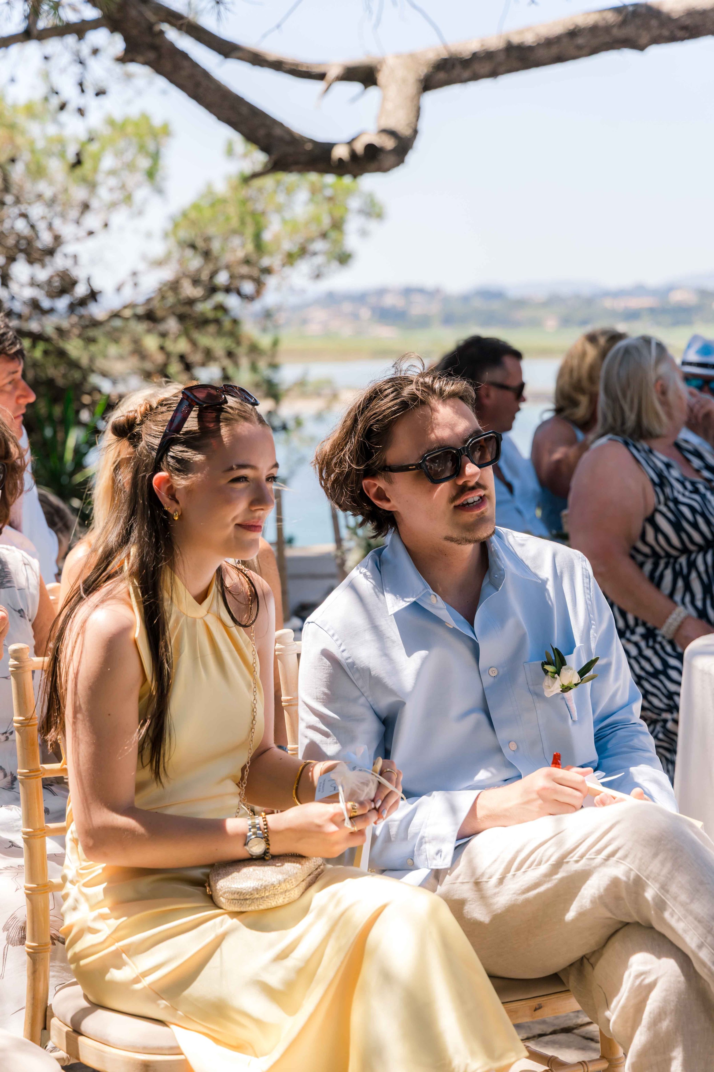 People sitting outdoors at a wedding ceremony with trees and water in the background.