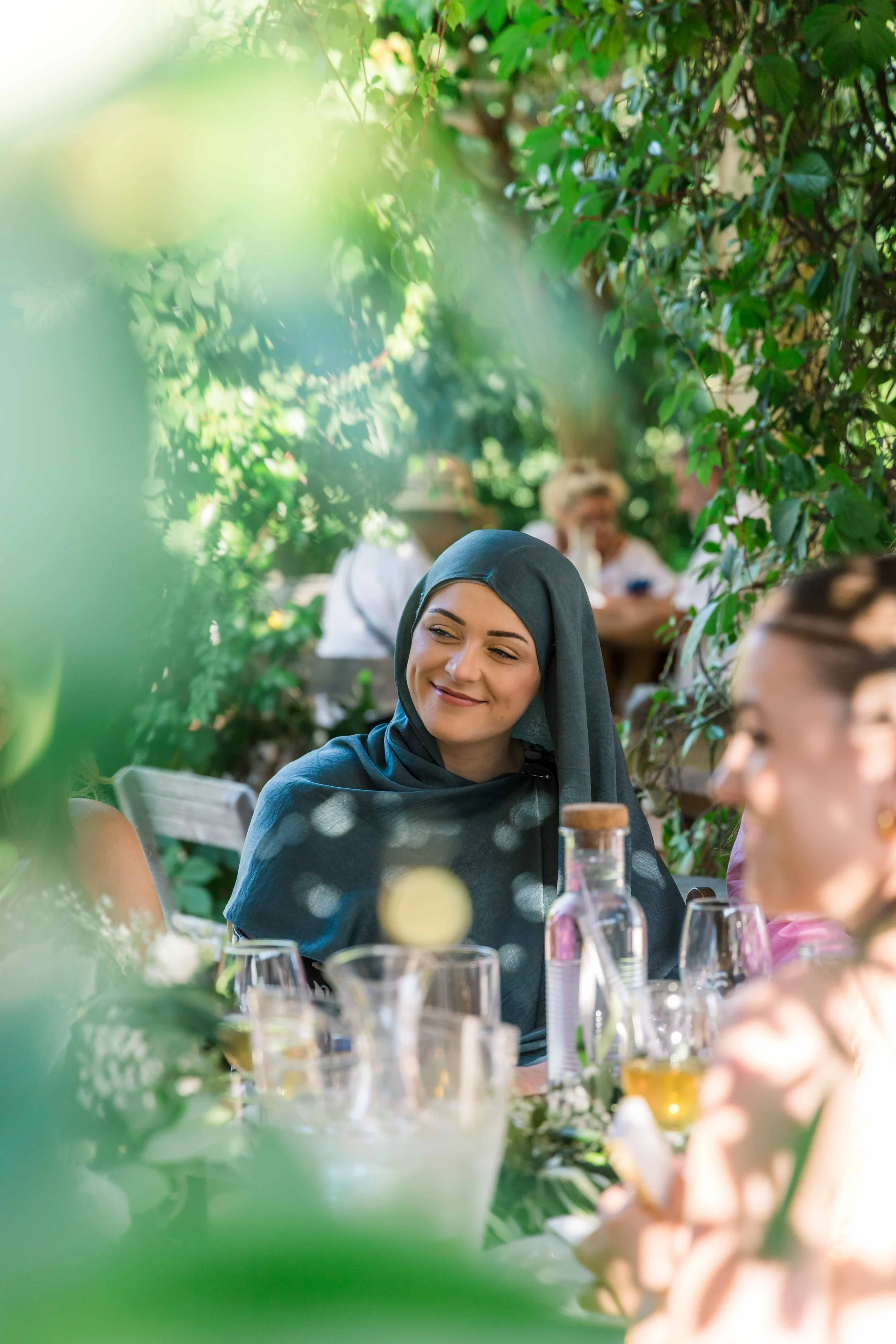 A woman smiling at a gathering or celebration, seated at a table outdoors, surrounded by greenery, with drinks and bottles visible on the table.