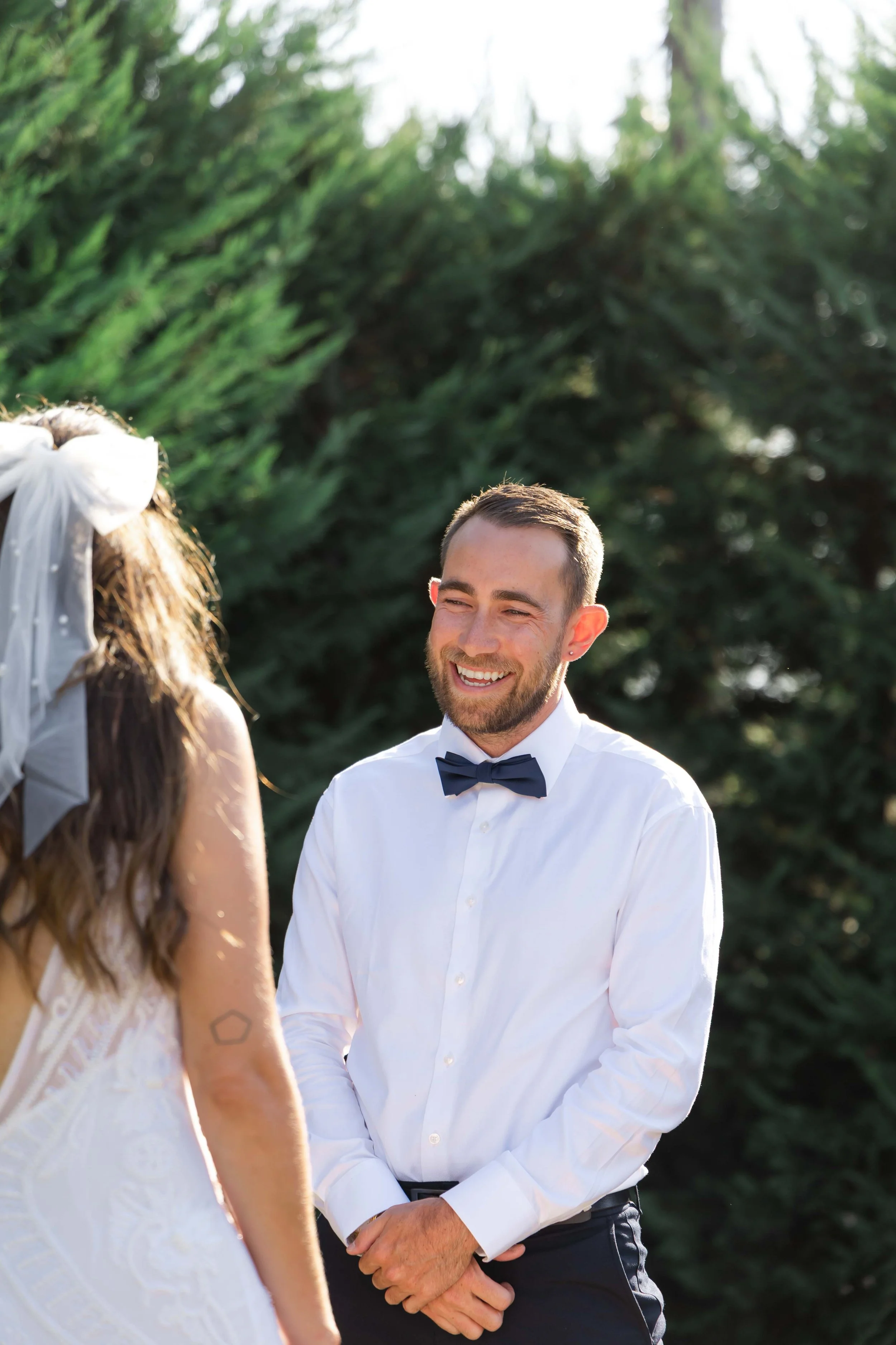 A man with a beard wearing a white dress shirt and a black bow tie is smiling at a woman during a wedding ceremony outdoors. The woman, seen from behind, has curly hair adorned with white ribbons and is wearing a white wedding dress, with only part o