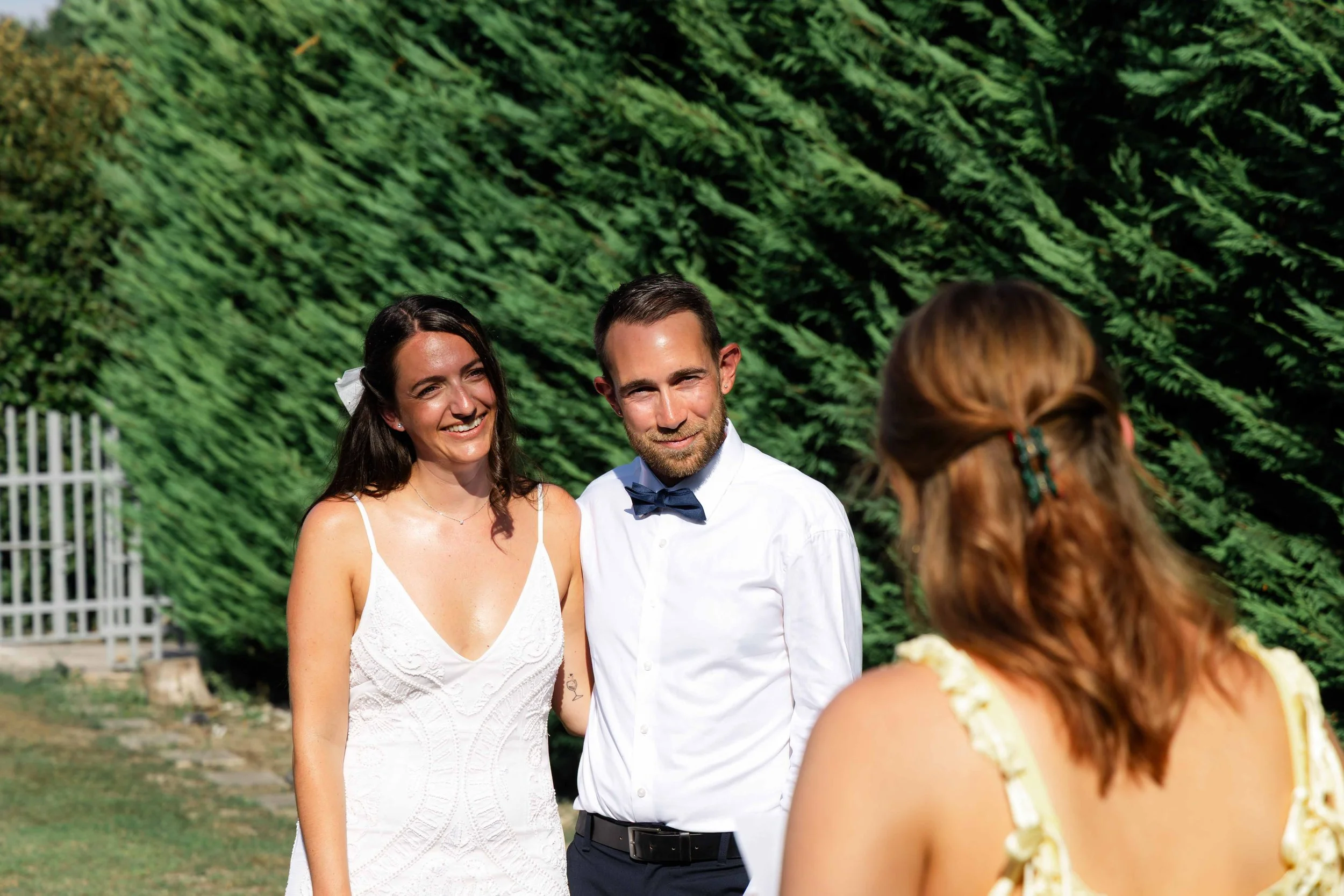 A bride and groom standing outdoors during a wedding ceremony, with greenery in the background. The bride has dark hair and is wearing a white dress, while the groom has short dark hair and is wearing a white shirt with a bow tie. Another person with