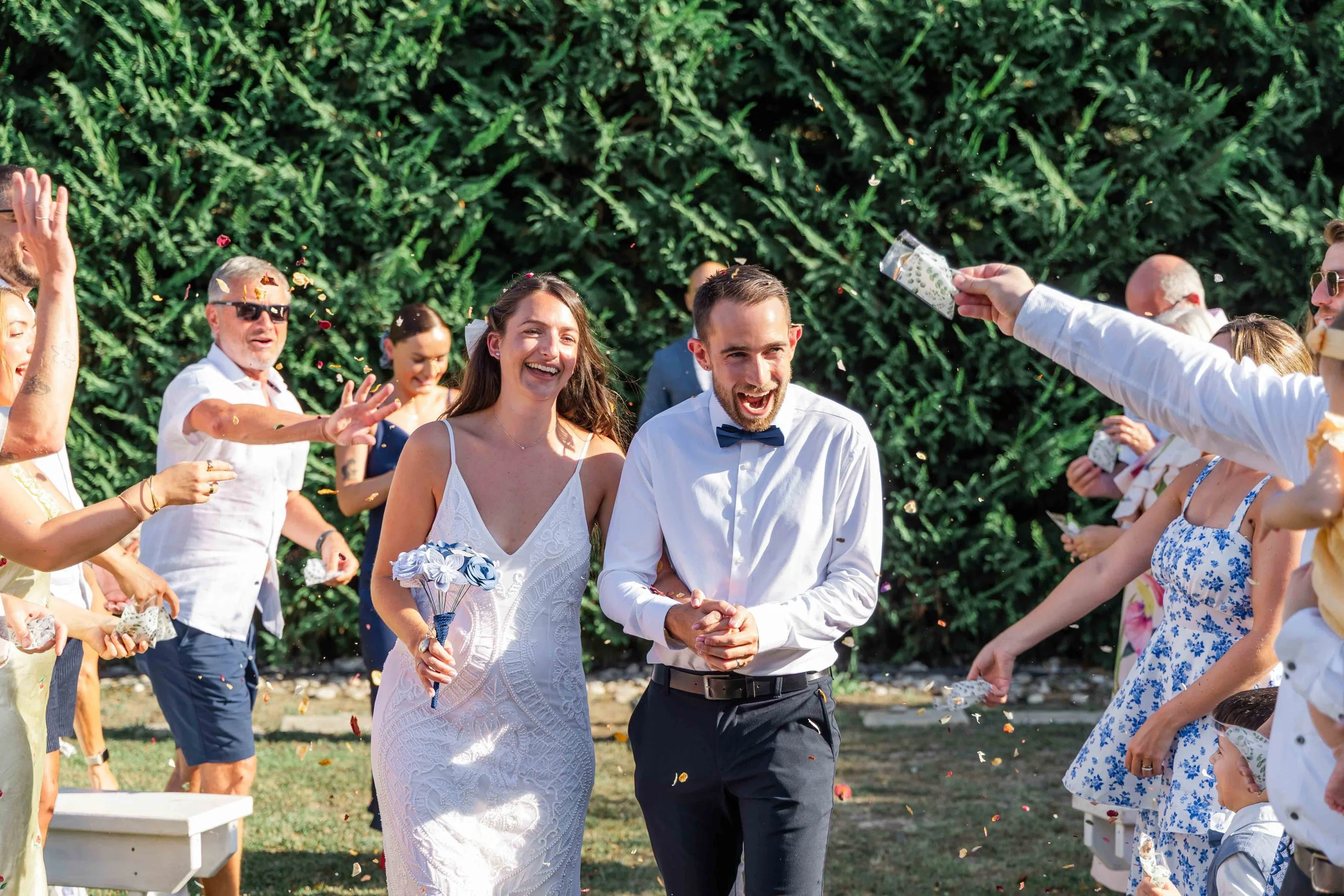 A bride and groom holding hands and smiling during a wedding celebration outdoors, surrounded by guests throwing flower petals.