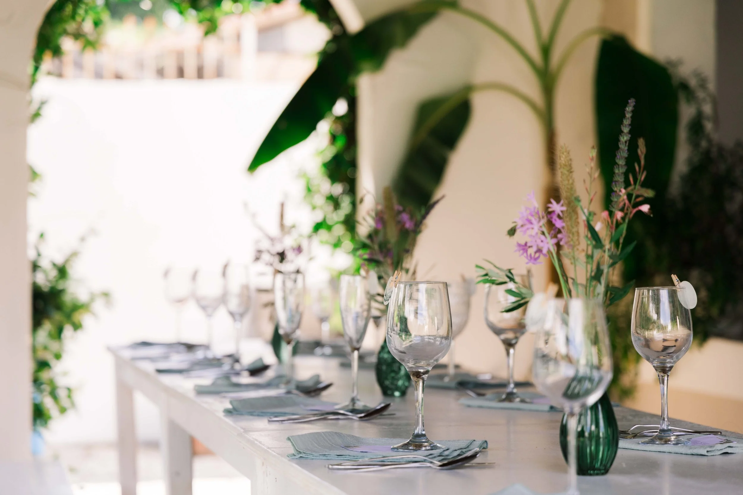 A table set for a meal with multiple wine glasses and a floral centerpiece in a green vase, with sunlight streaming through a window.