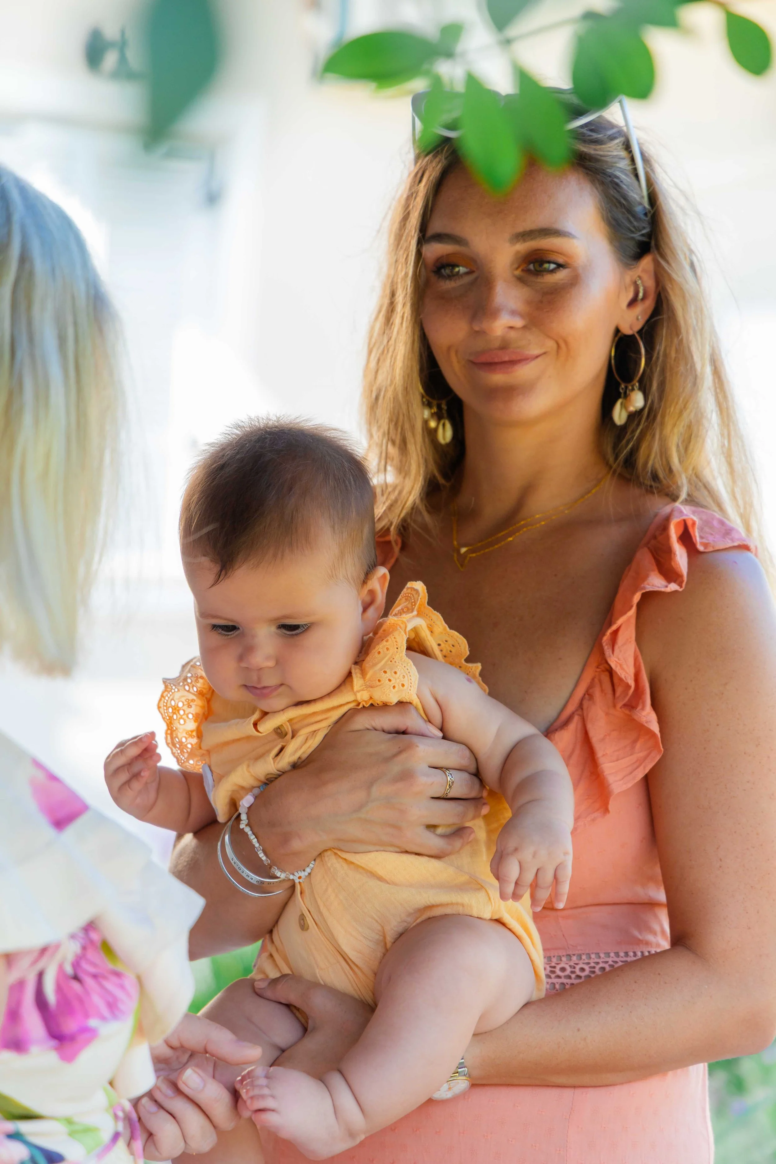 A woman with light brown, wavy hair holding a young child in her arms, both wearing orange outfits. The woman has earrings and a necklace, and appears to be smiling softly. They are outdoors, with green foliage and colorful decorations in the backgro