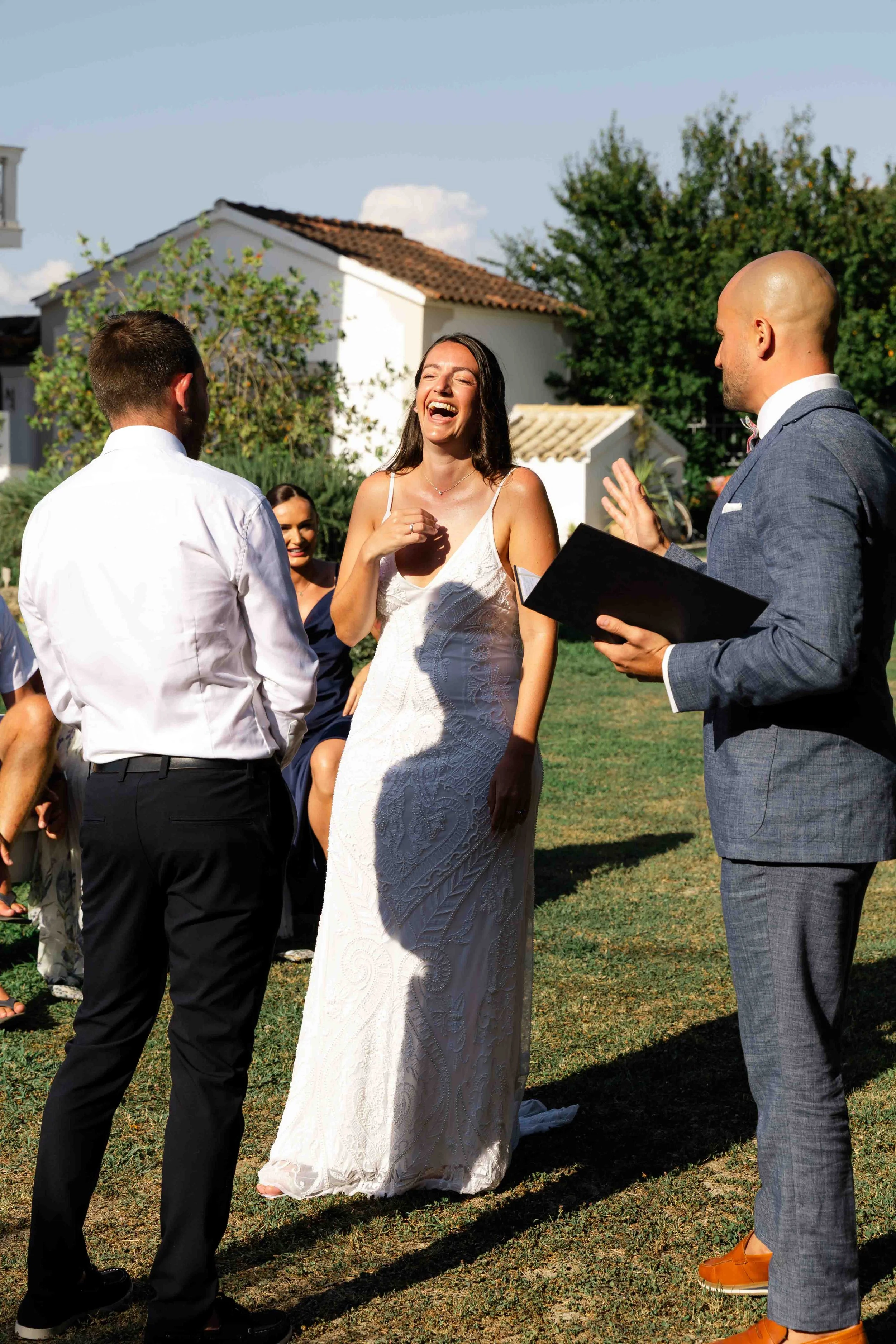 A woman in a white wedding dress and a man in a gray suit with orange shoes standing outdoors, smiling and interacting with others at a celebration.