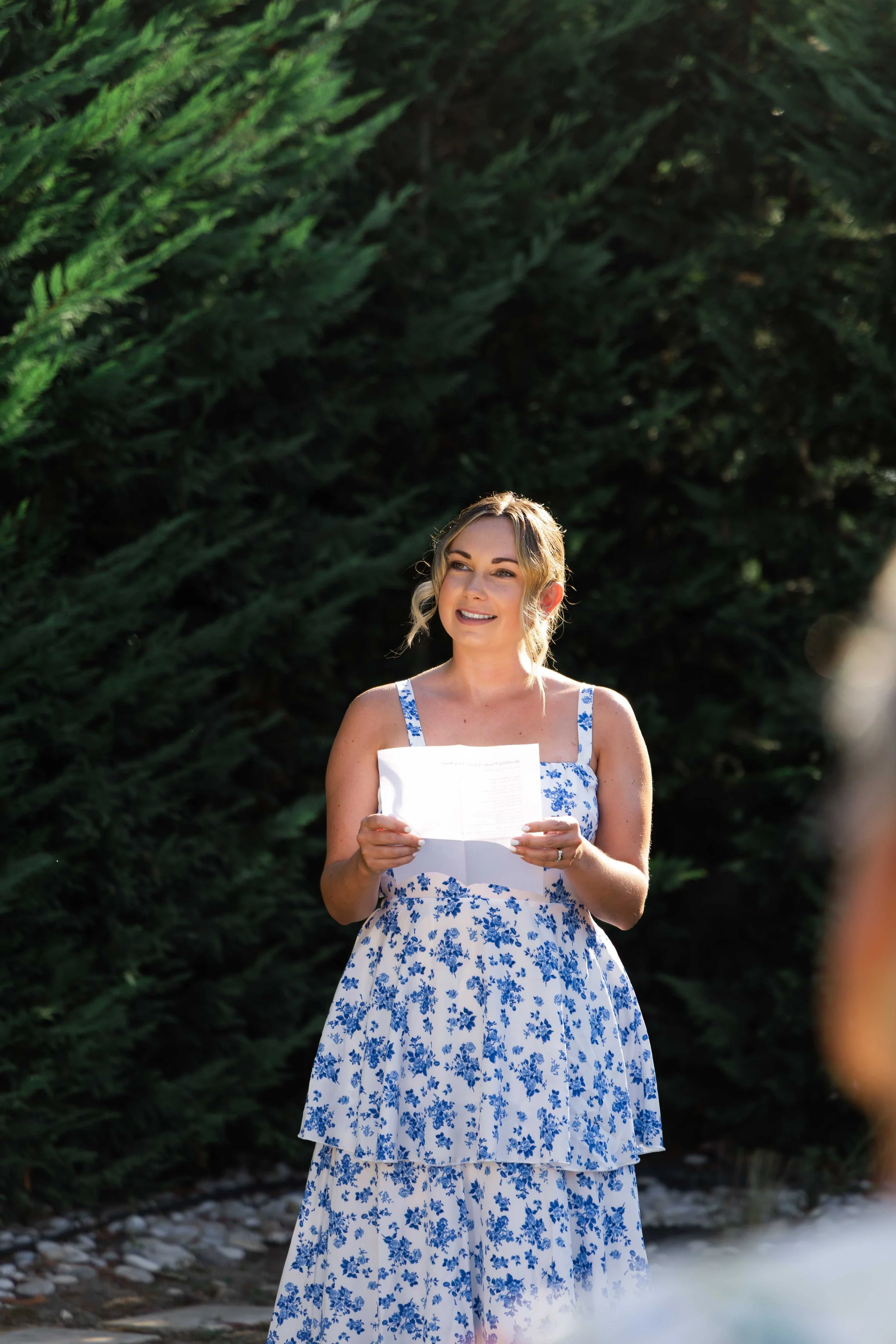 A woman in a white and blue floral dress holding a piece of paper, standing outdoors in front of green bushes, smiling.