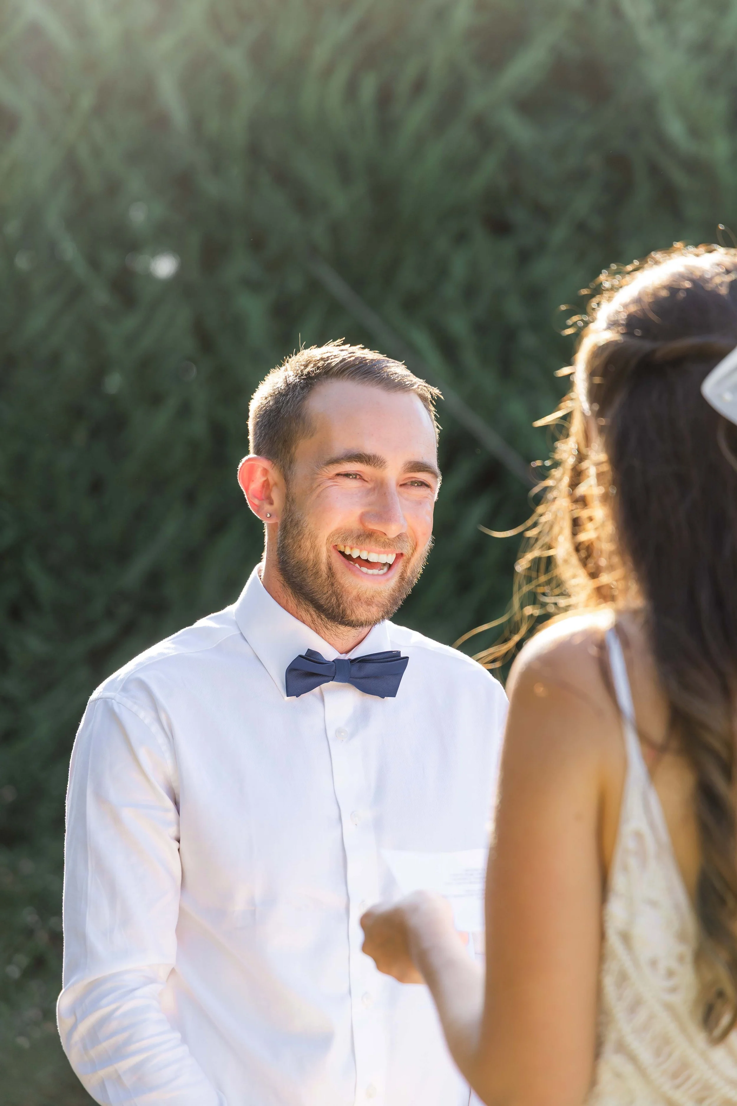 A man wearing a white dress shirt and bow tie smiling at an outdoor event, with a woman in a white dress with lace details partially visible in the foreground.