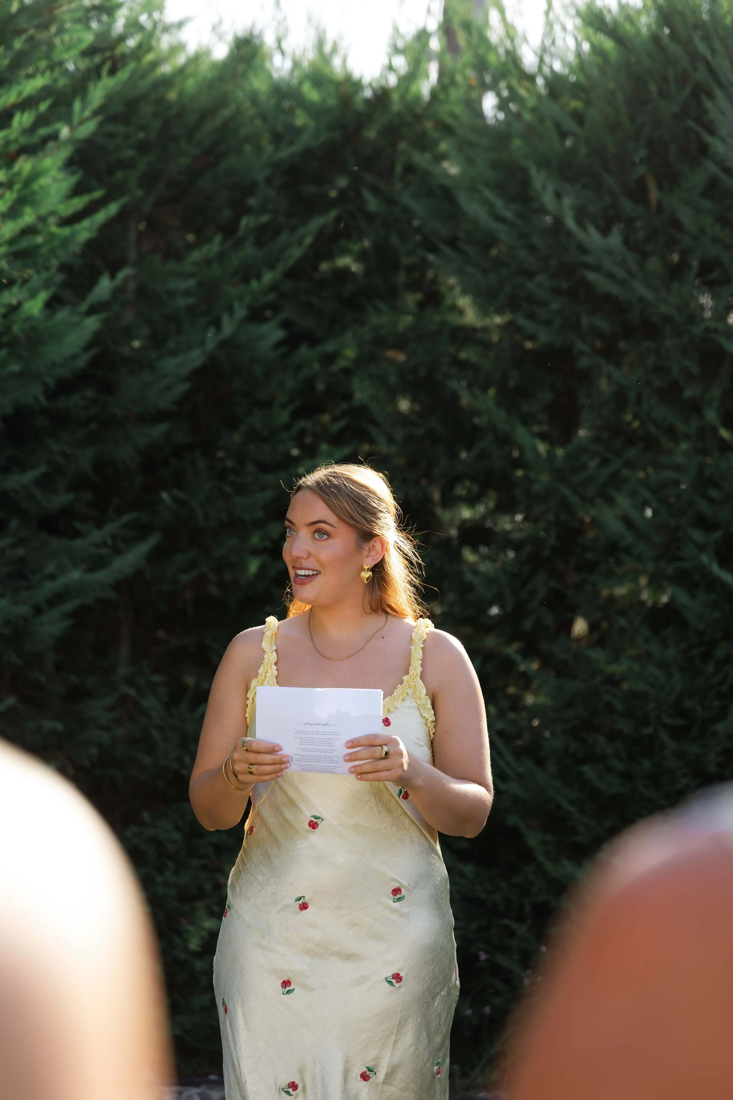 A woman in a yellow dress with red cherries holds a piece of paper. She has blonde hair, heart-shaped earrings, and is outdoors with green trees in the background.