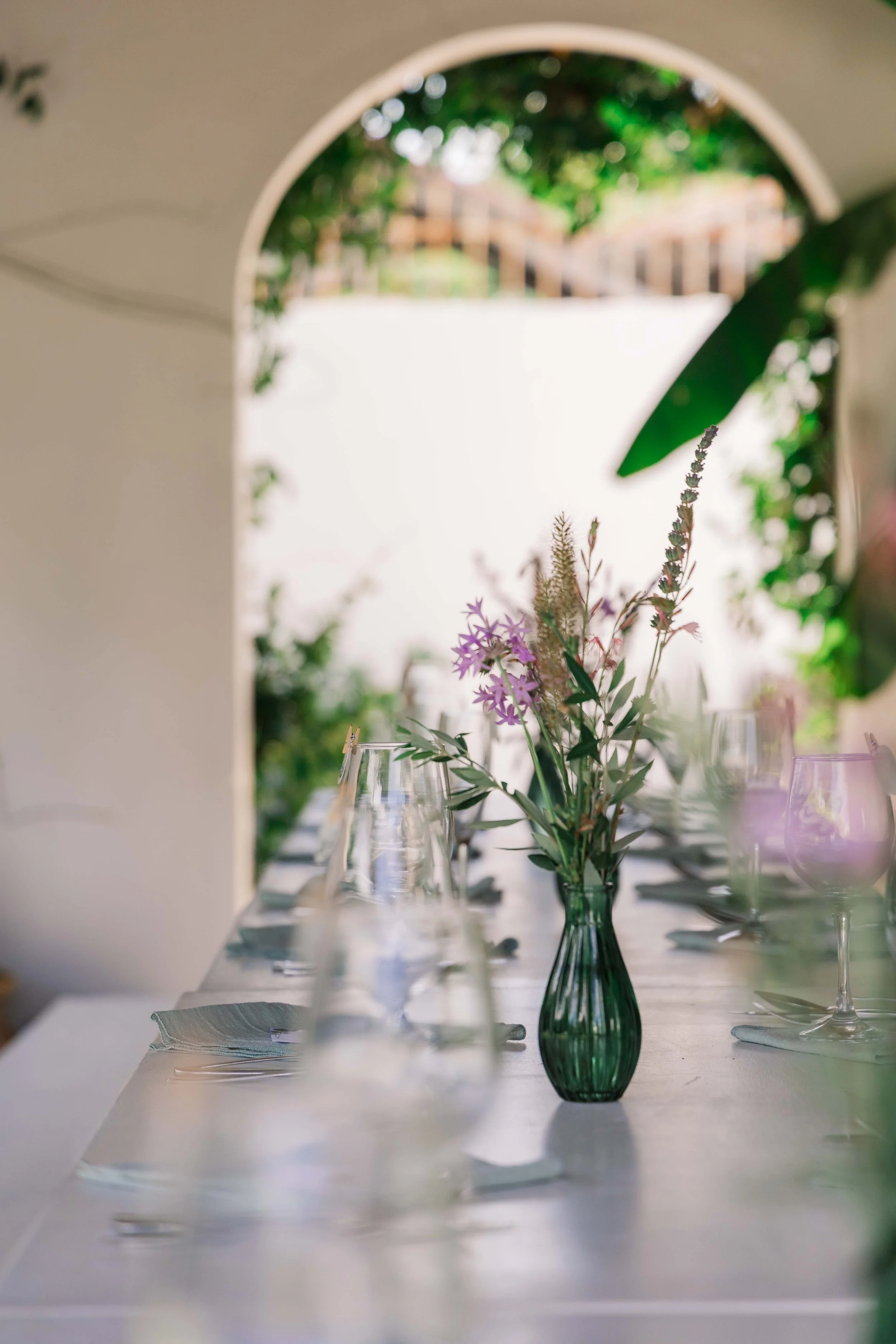 A close-up of a table with a green glass vase holding pink and purple flowers, set with glassware and cloth napkins in an outdoor setting with a garden view through an arched opening.