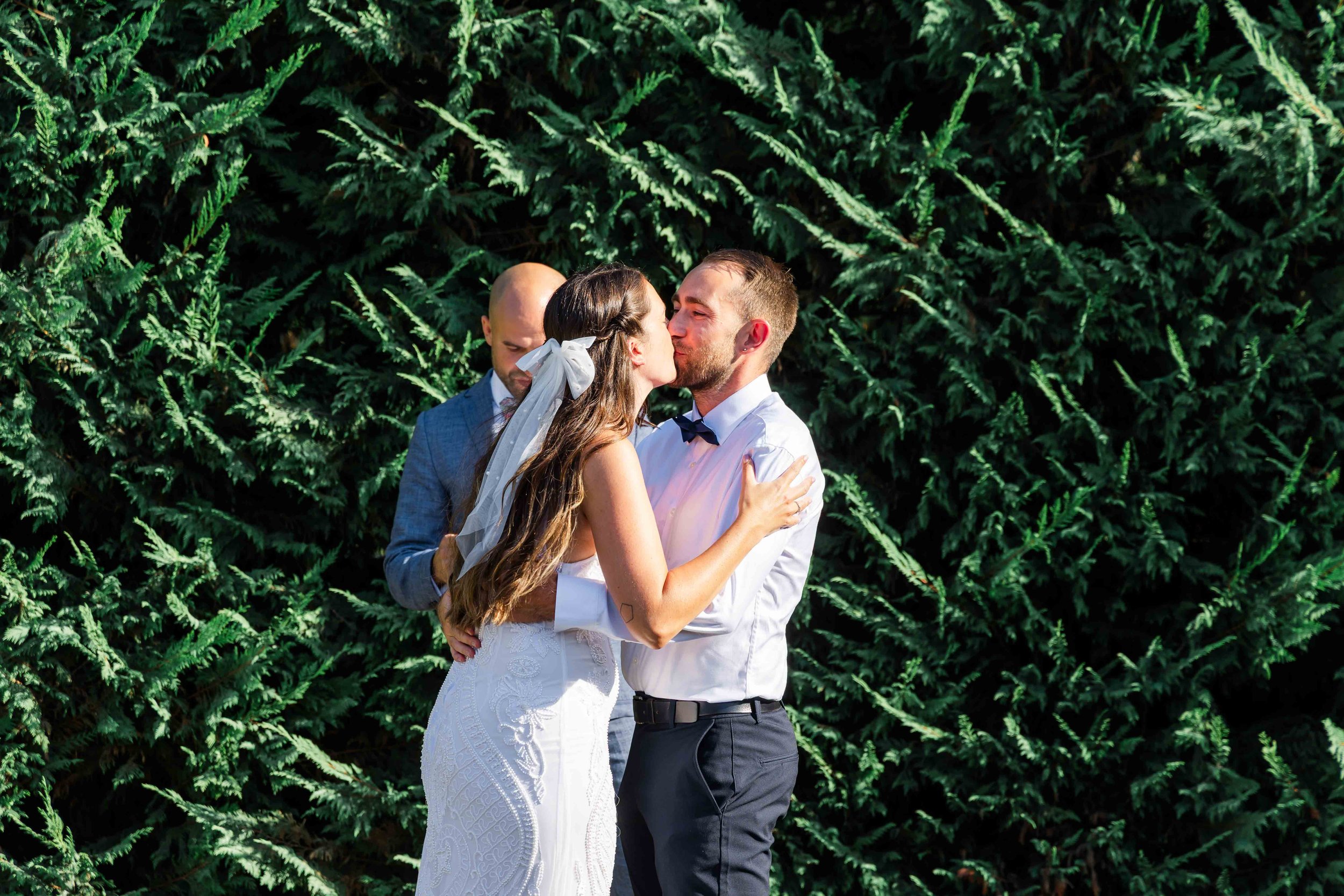 A couple getting married, kissing, with a wedding officiant in the background, outdoors in front of lush green foliage.