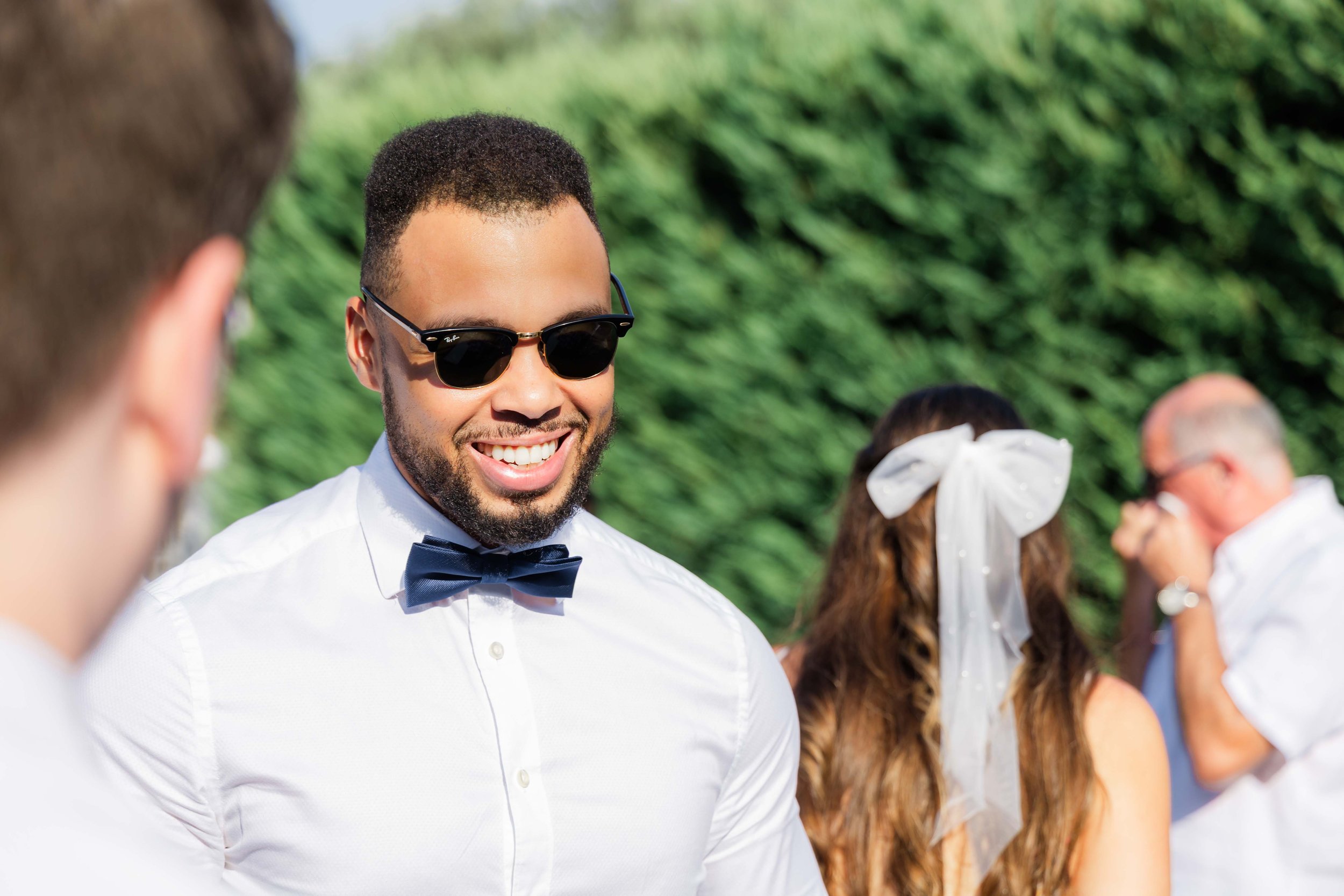 Smiling man wearing sunglasses, white shirt, and bow tie during an outdoor event. Other people, including woman with a white bow in her hair, are visible in the background.