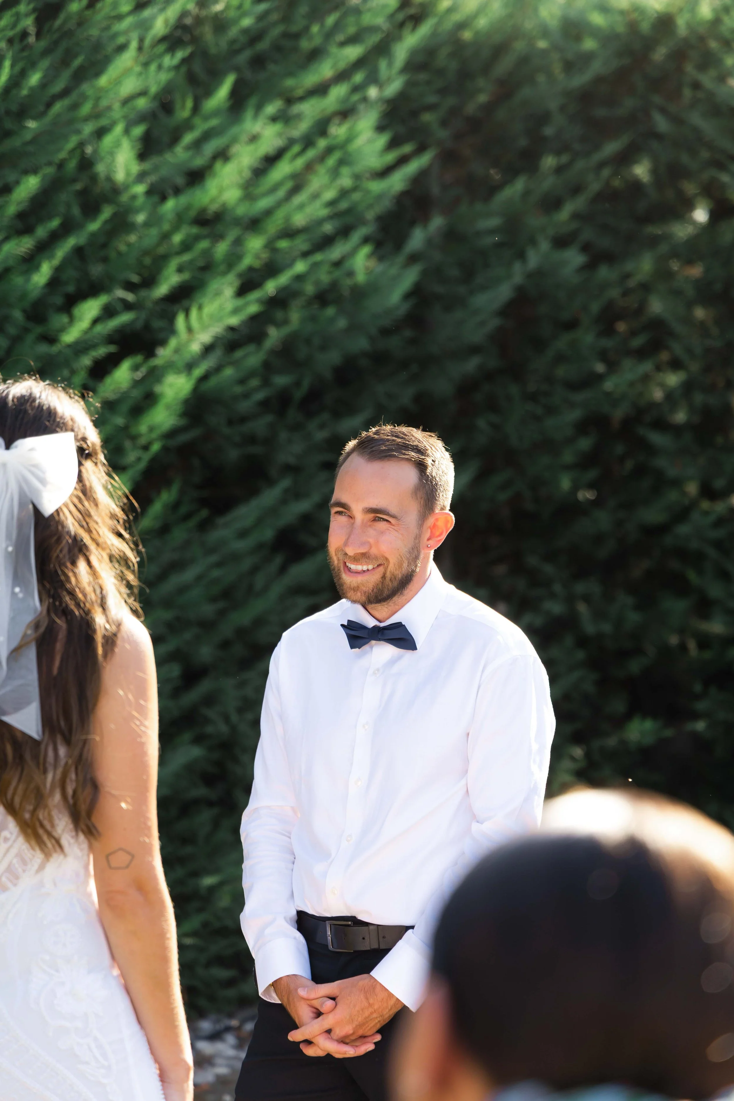 A man with a beard and short hair, dressed in a white shirt and black bow tie, smiling and holding his hands together in front of him during an outdoor event.