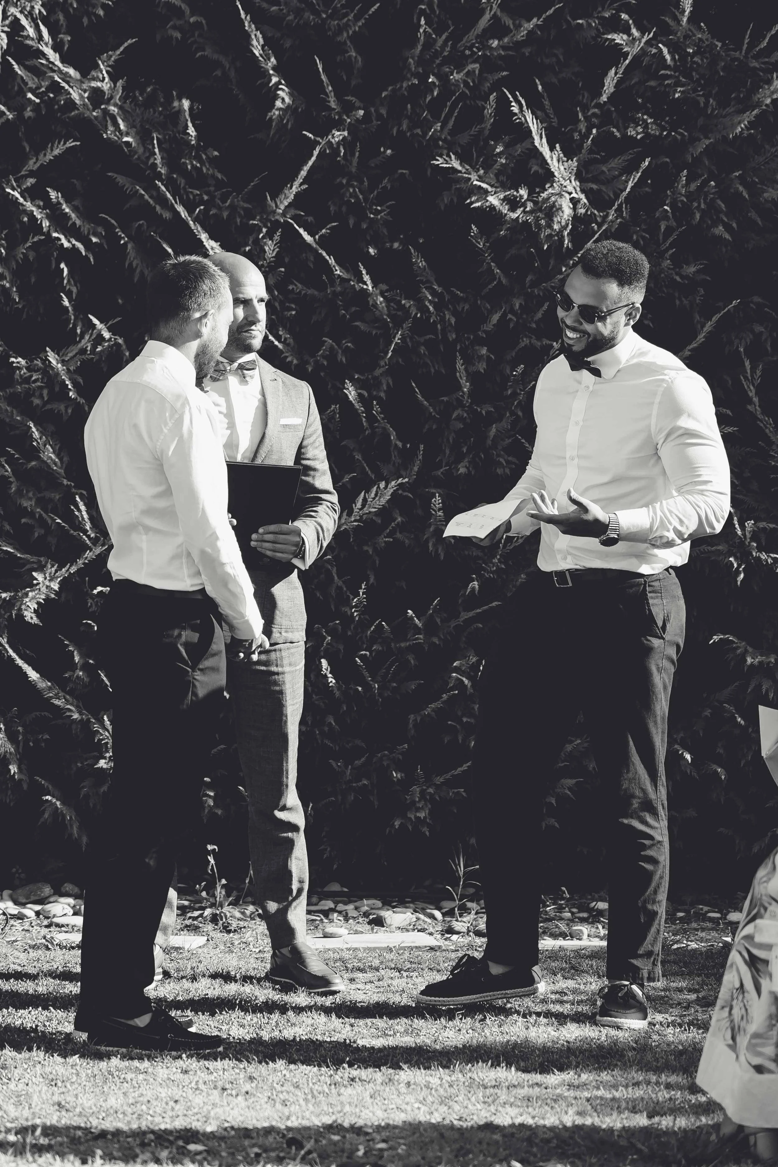 Four men at an outdoor wedding ceremony, three of them engaged in conversation, with one holding a certificate, against a background of dense trees.
