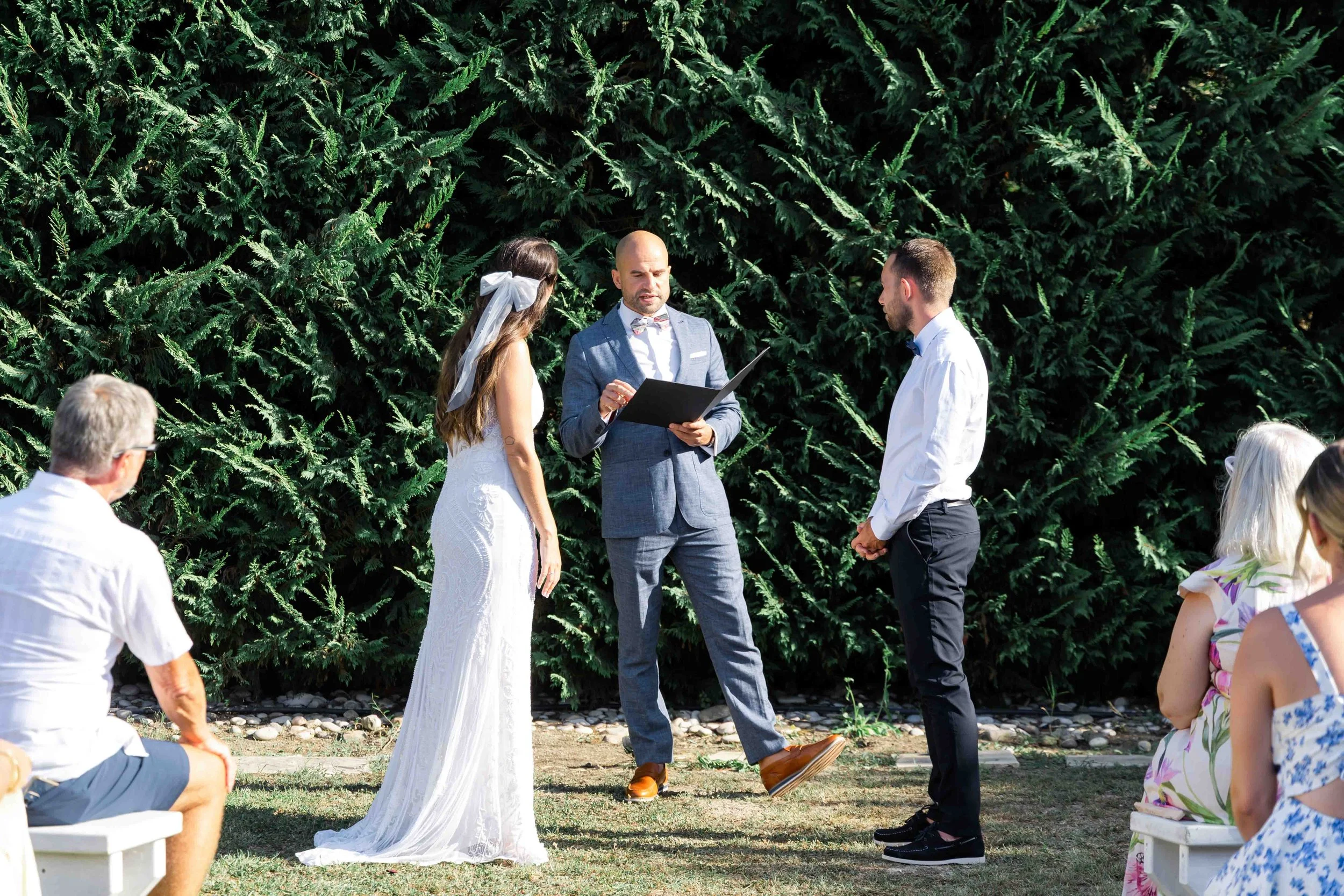A wedding ceremony outdoors with a bride, groom, and officiant standing in front of a green hedge. The bride is in a white dress with a bow in her hair. The officiant is in a light gray suit. Guests are seated on white benches, with some guests visib