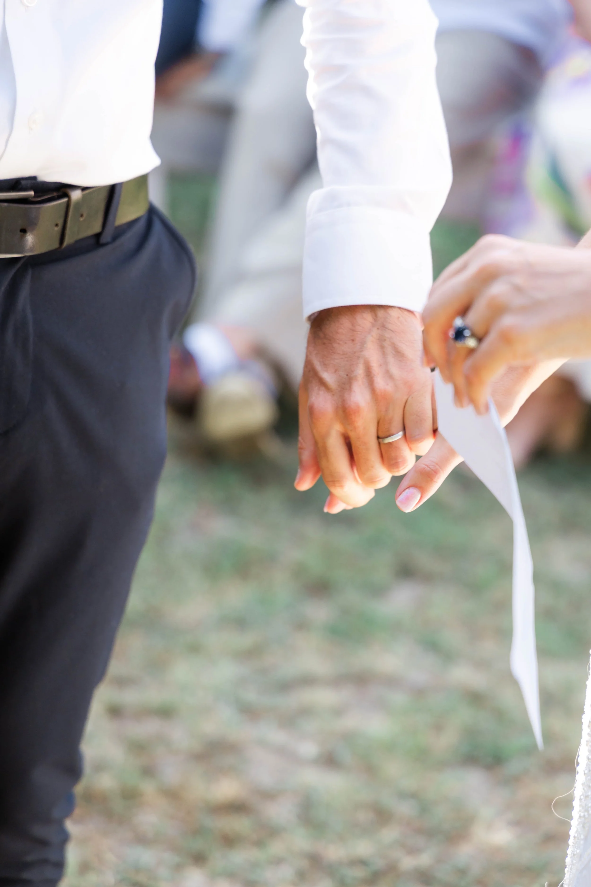 Close-up of two people holding hands, attending a wedding ceremony outdoors, with one wearing a wedding ring.