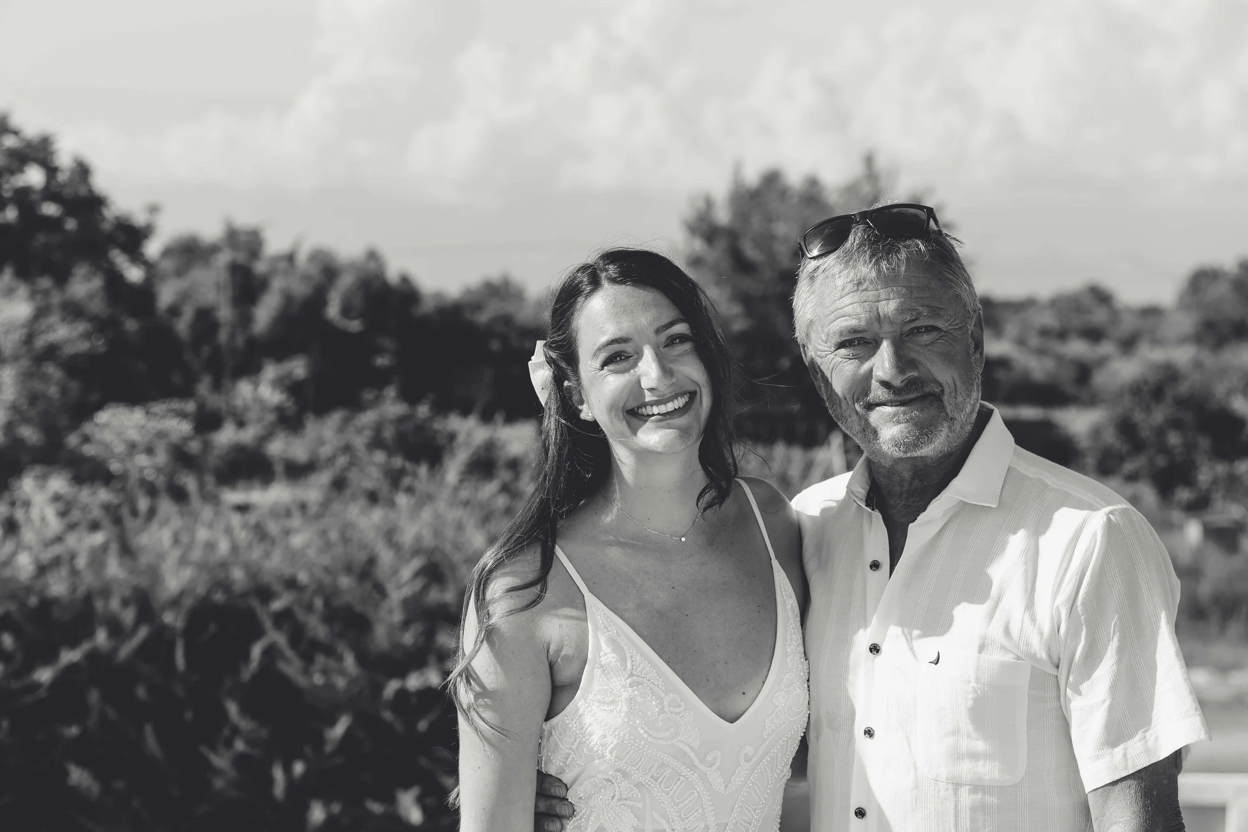 A smiling young woman and an older man standing close together outdoors, both dressed in summer attire, with trees and a cloudy sky in the background.