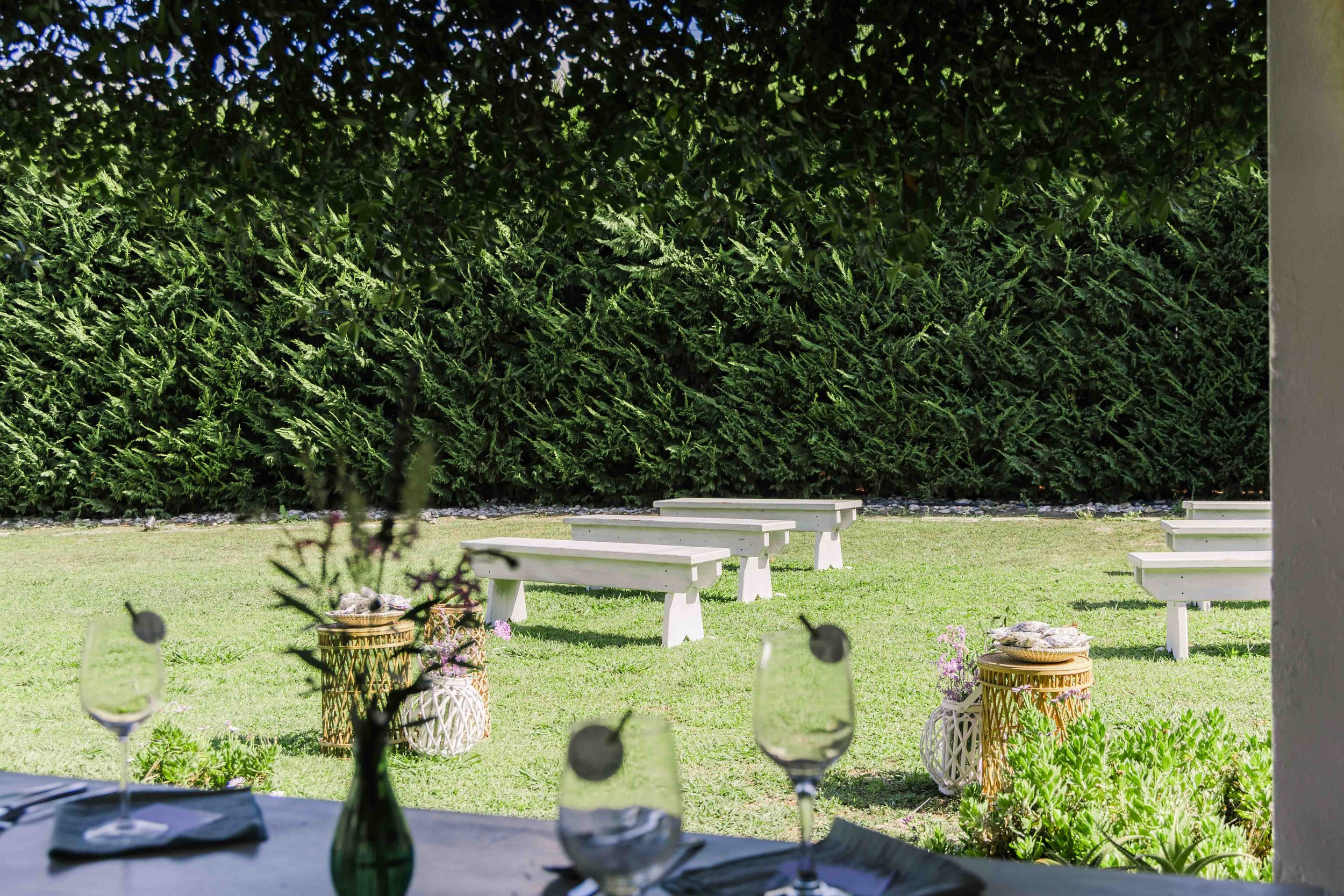 Outdoor venue with tables and glasses on a table in the foreground, and benches with a tall green hedge in the background.