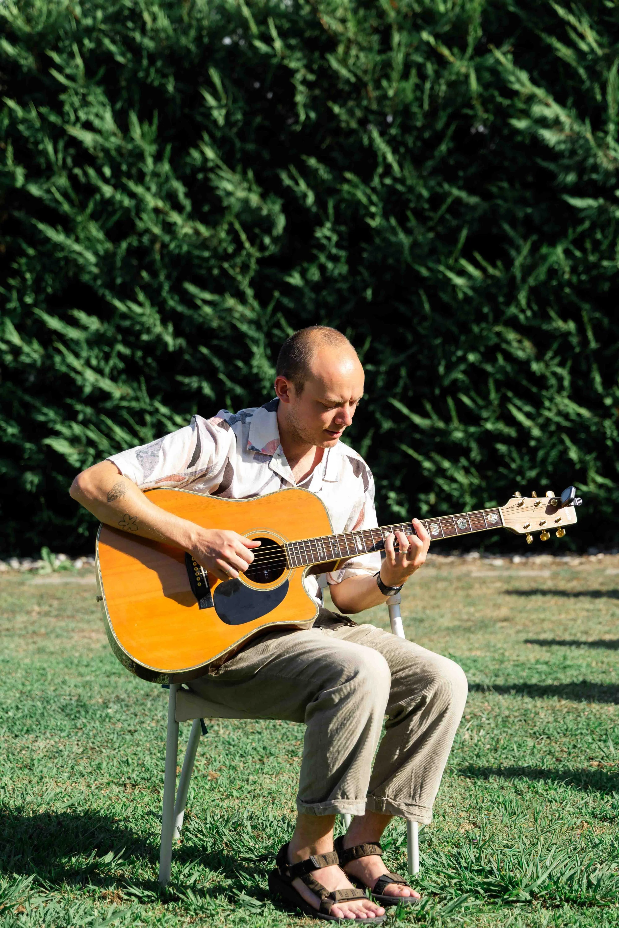A man with a shaved head and tattoos on his arm, wearing a short-sleeved button-up shirt and beige shorts, is sitting outdoors on a white chair, playing an acoustic guitar on a sunny day with green grass and bushes in the background.