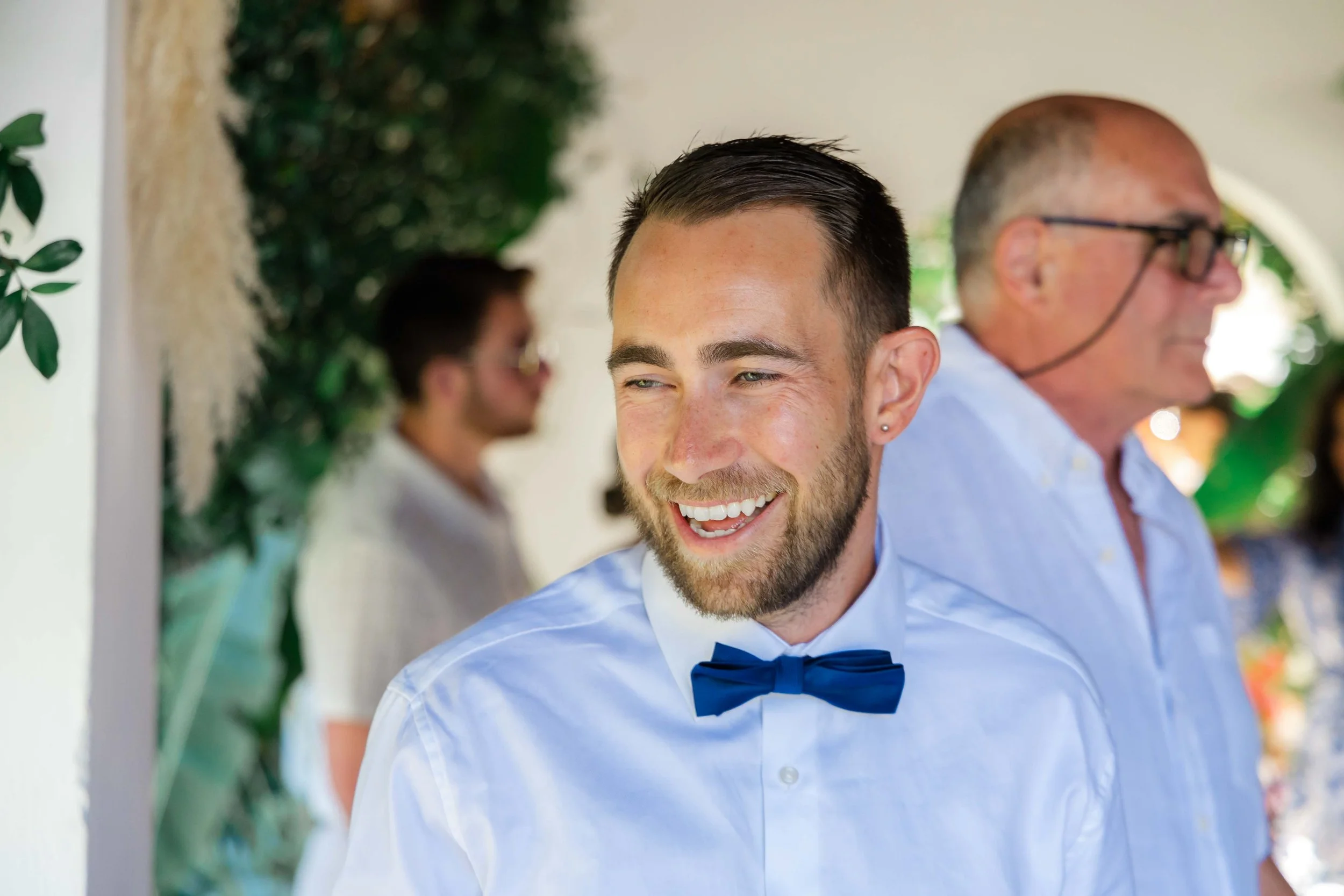 A young man with short dark hair, a beard, and an earring, smiling while wearing a white shirt and blue bow tie. In the background, an older man with glasses and a receding hairline is also smiling, with a woman and greenery partially visible.