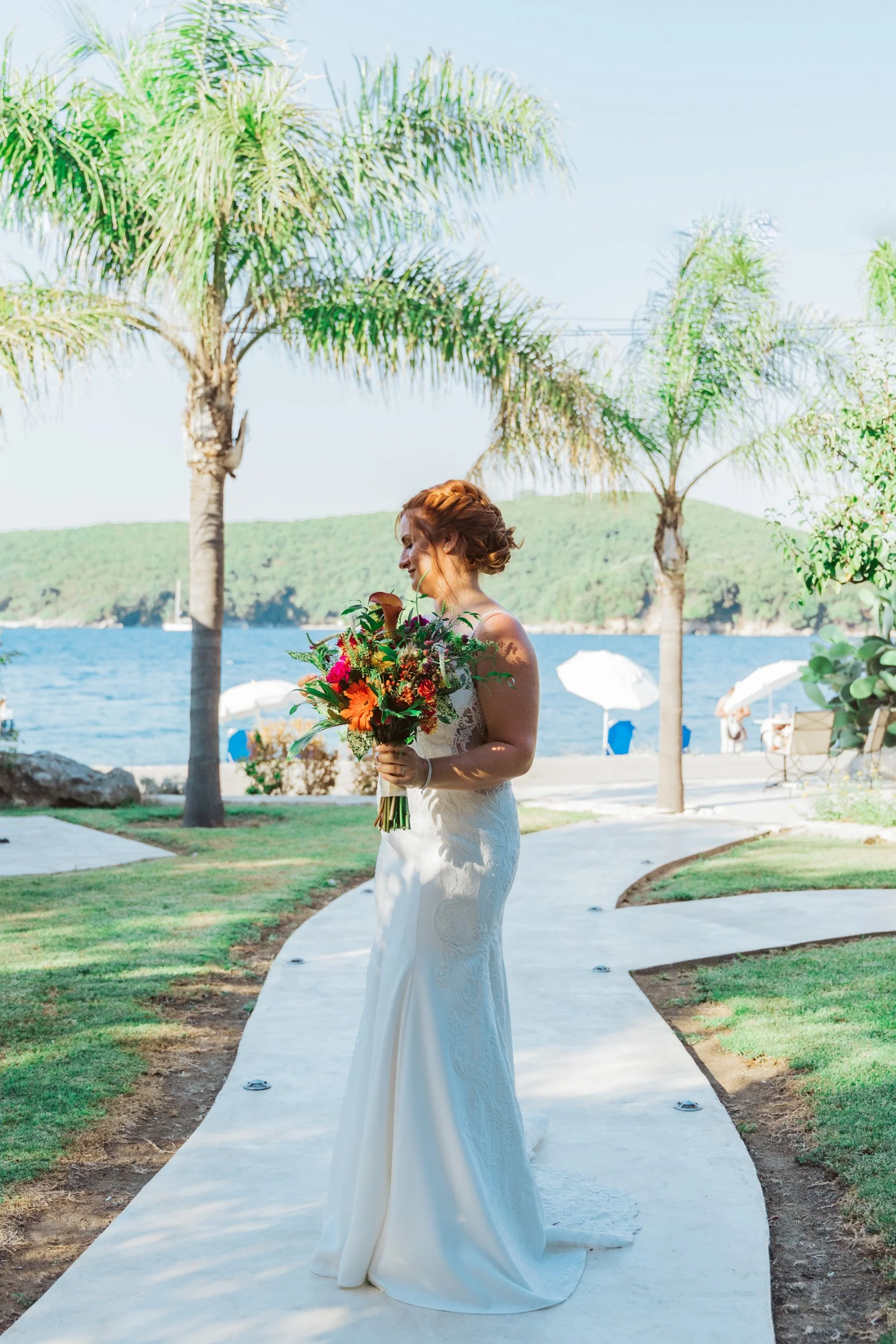 A bride holding a colorful bouquet of flowers on a winding pathway near a body of water, with palm trees and a green hillside in the background during daytime.