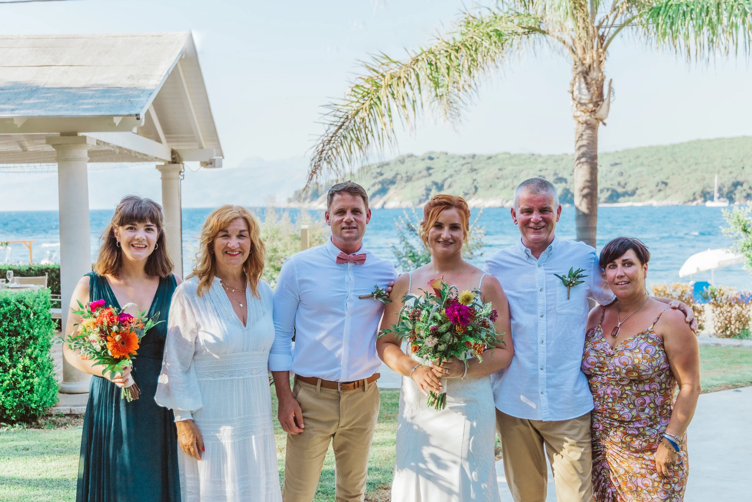 Group of six people, including a bride and groom, smiling outdoors near water with a sailboat, palm tree, and hillside in the background.